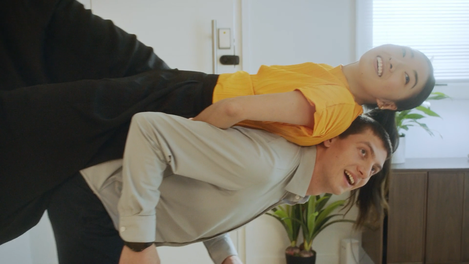 Two people, a woman in a yellow top and a man in a beige shirt, are smiling and leaning close together in an office setting with a plant and window in the background.