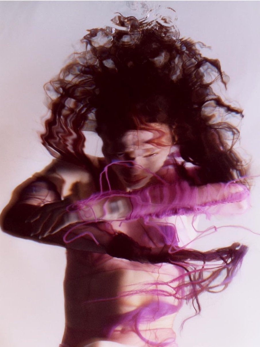 A woman with curly hair is lying with her hand behind her head, with pink and purple light streaks across her face and body, creating a surreal, abstract effect.