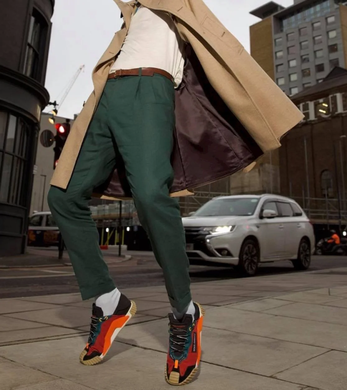 Person wearing a tan trench coat, white shirt, green pants, and colorful sneakers walking on city sidewalk with cars and buildings in background.