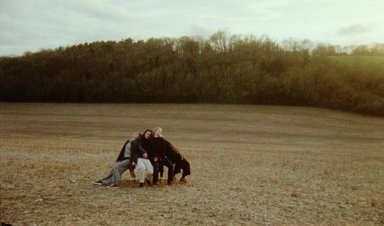 Four people sitting on a bench in an open field with a wooded hillside in the background.