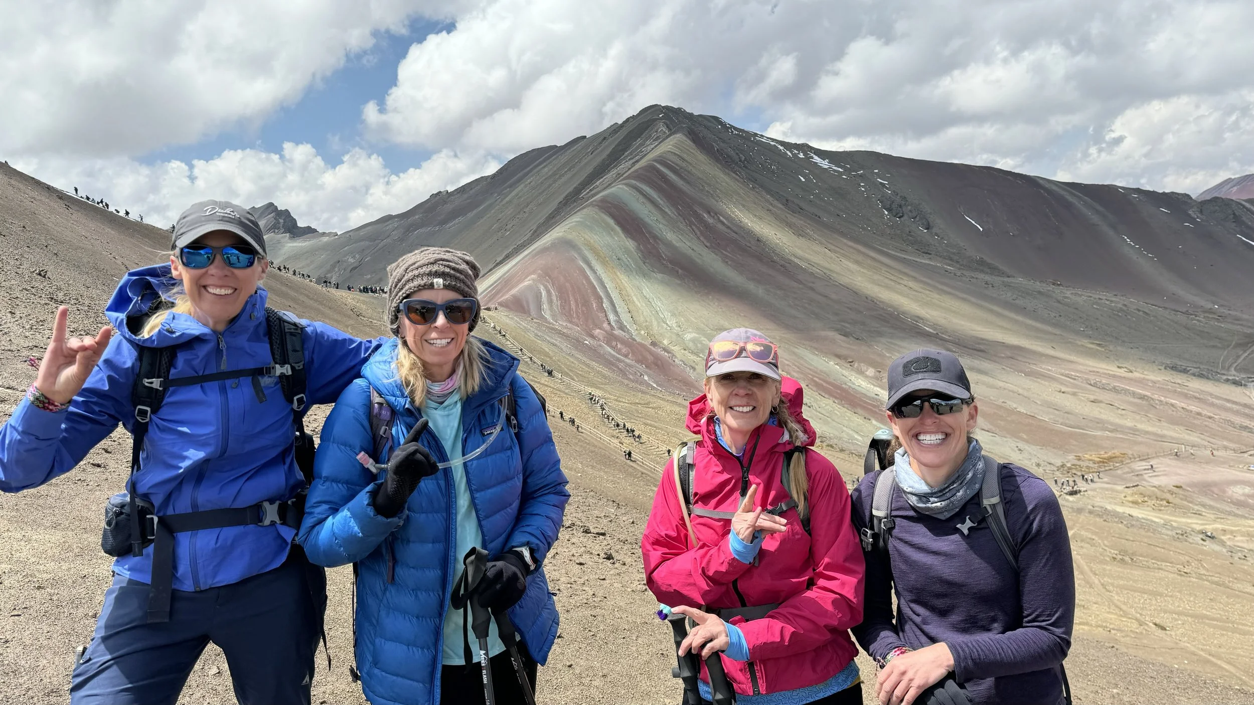 Four women hiking on a mountain trail with colorful volcanic mountain in the background, smiling and posing for a photo.