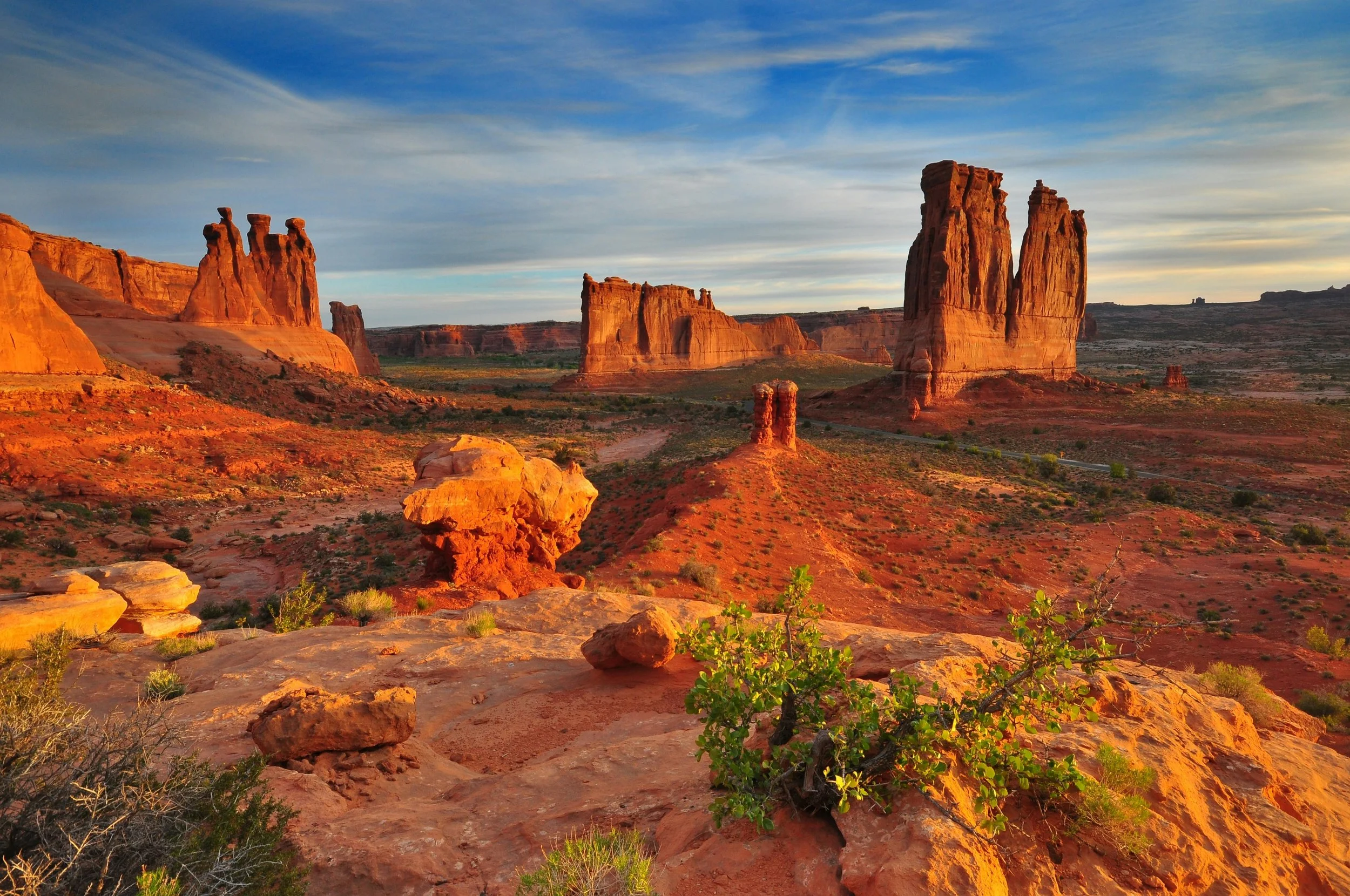 Sunset over red rock formations in a desert landscape with sparse green vegetation, blue sky, and wispy clouds.