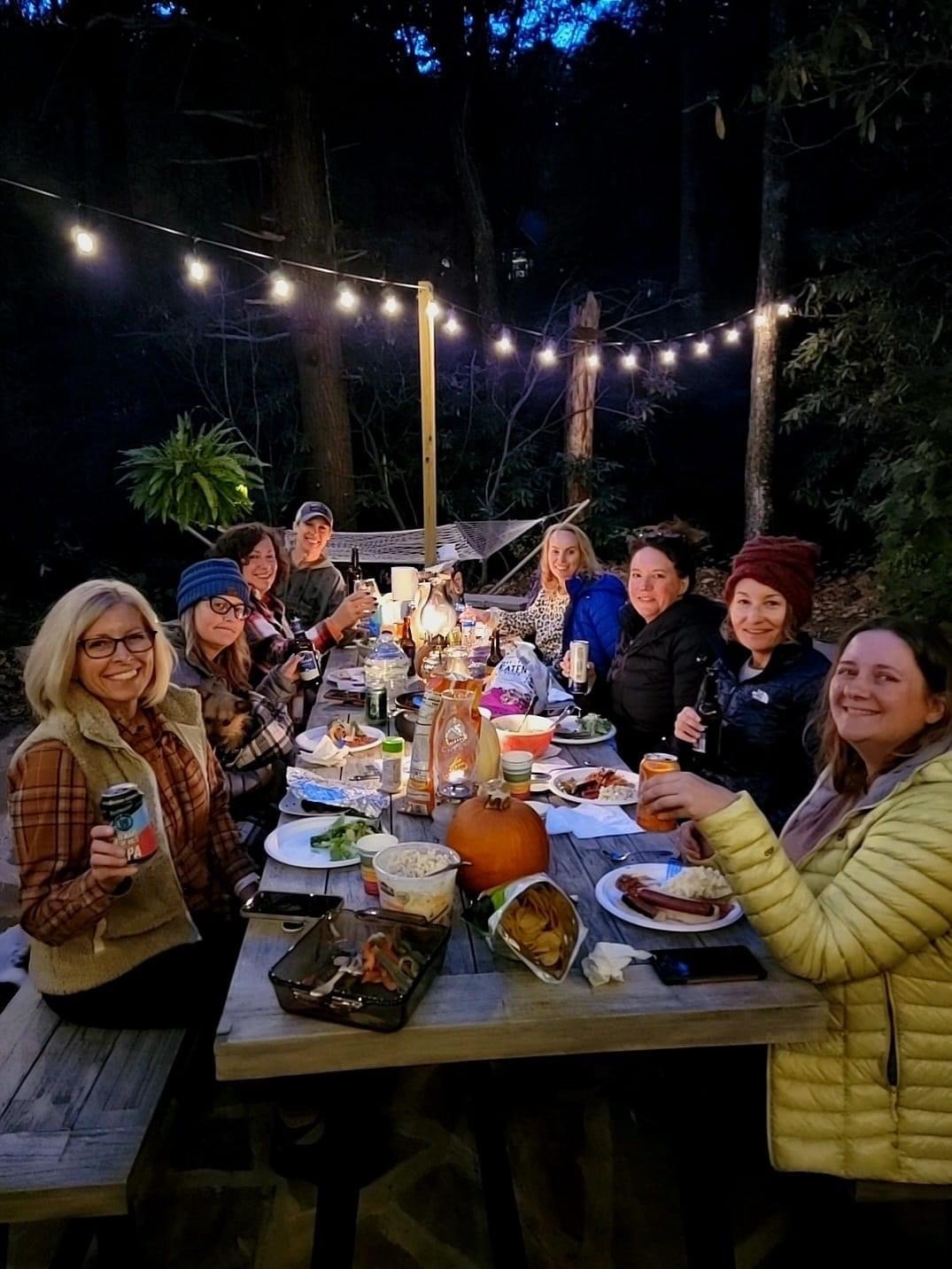 Group of people sitting at a long outdoor dinner table under string lights in a wooded area, enjoying food and drinks.