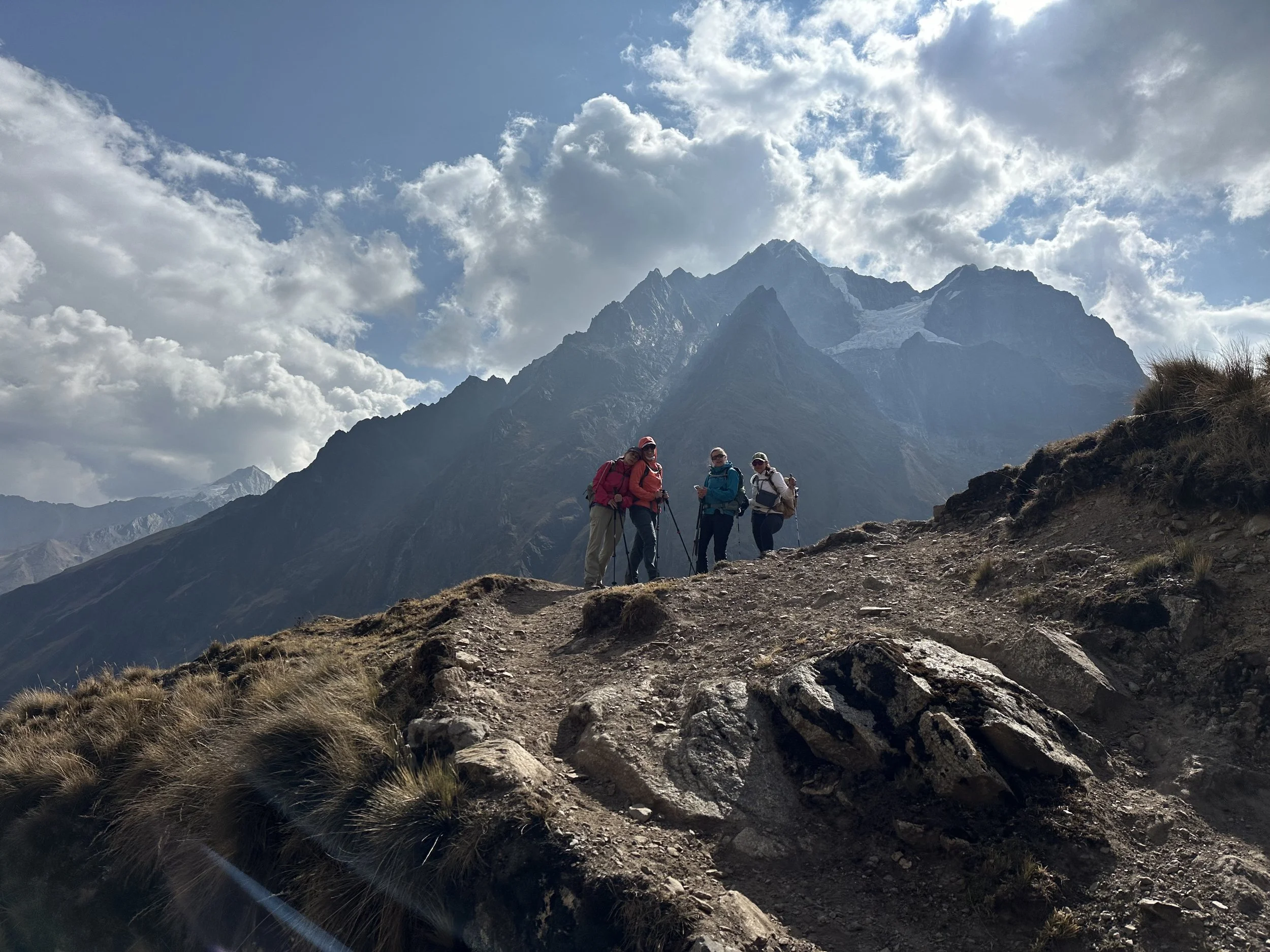 Group of hikers walking on a rocky mountain trail with a backdrop of tall snow-capped mountains and partly cloudy sky.