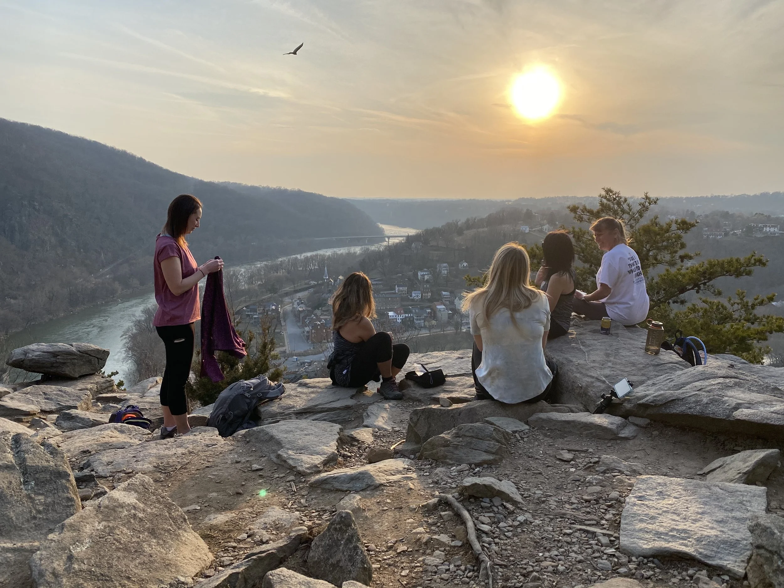 A group of people sitting and standing on rocky terrain a hillside during sunset, overlooking a river, bridge, and town below with houses and trees.