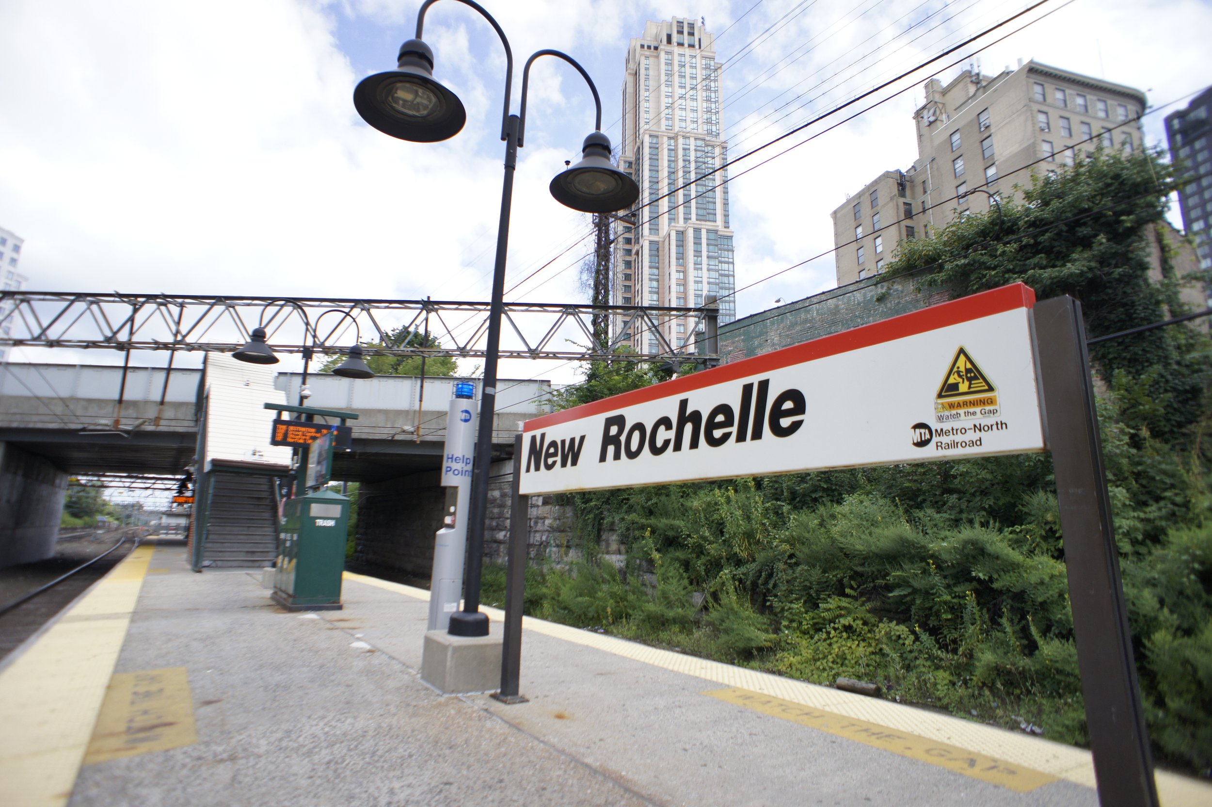 New Rochelle Metro-North Railroad station platform with a sign, street lamps, and train tracks, city buildings in the background, and overhead electrical wires.