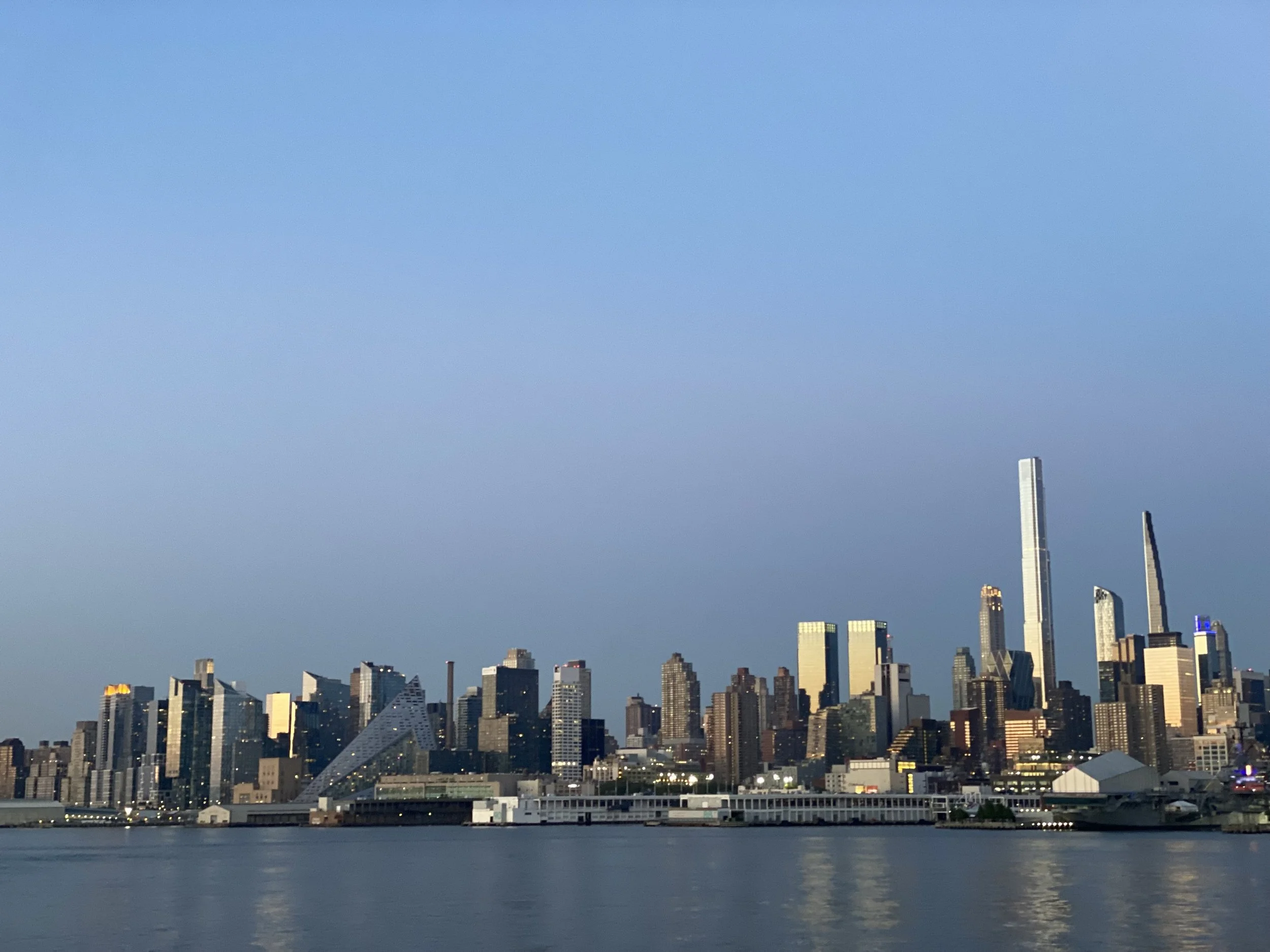 A midtown city skyline with tall skyscrapers and buildings along a waterfront, with a clear blue sky above.