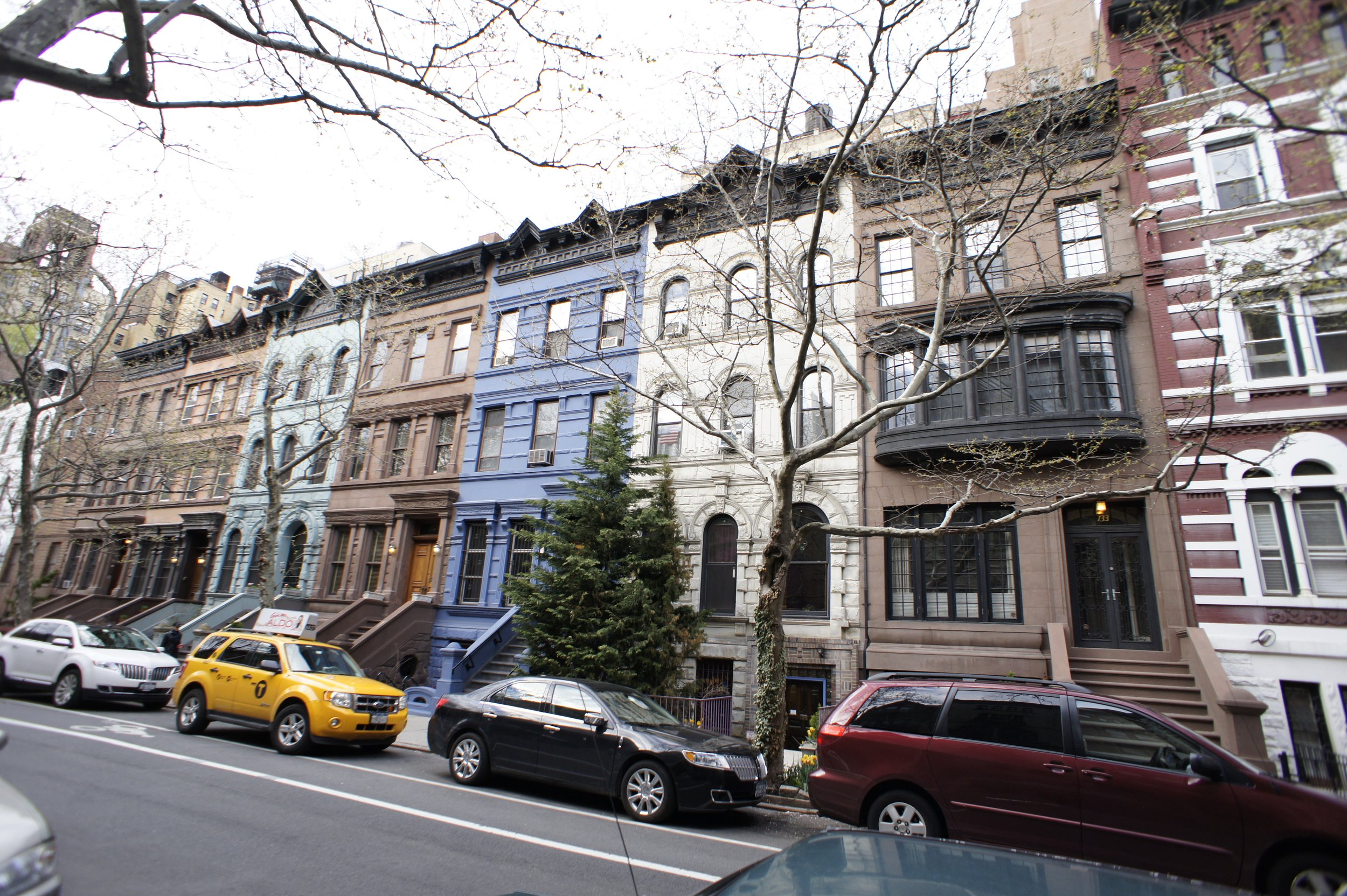 Row of colorful brownstone buildings with trees in front and parked cars on the street in Upper West Side, Manhattan.