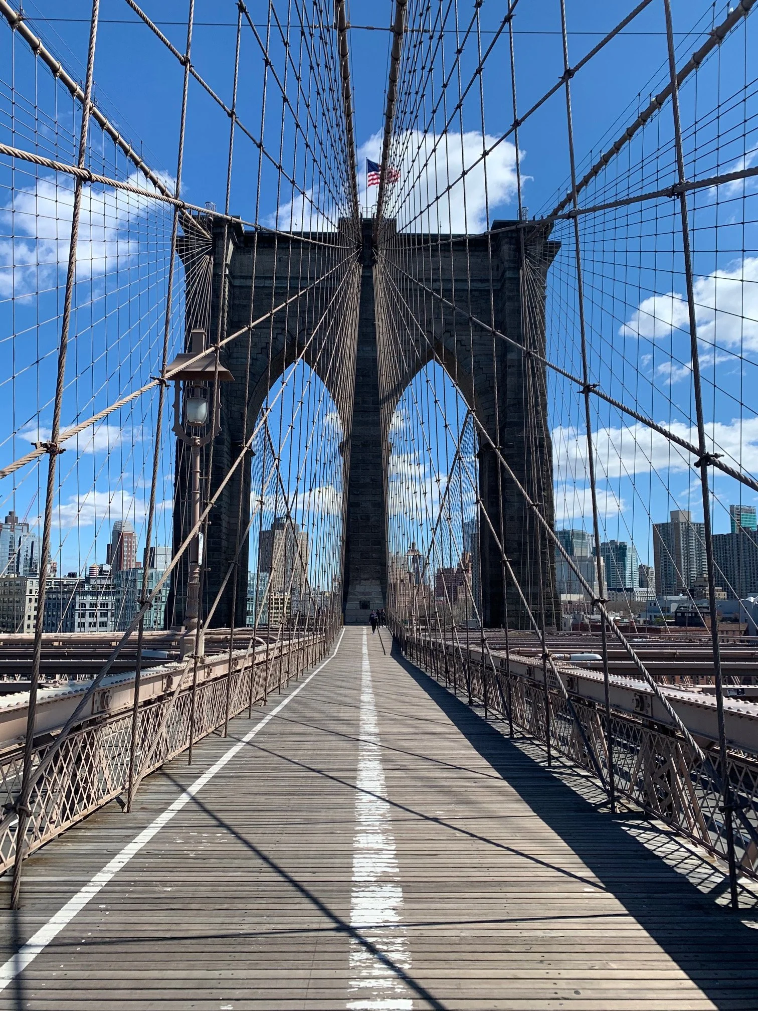 View from Brooklyn Bridge walkway showing the bridge's stone towers, cables, and a city skyline in the background under a clear blue sky.
