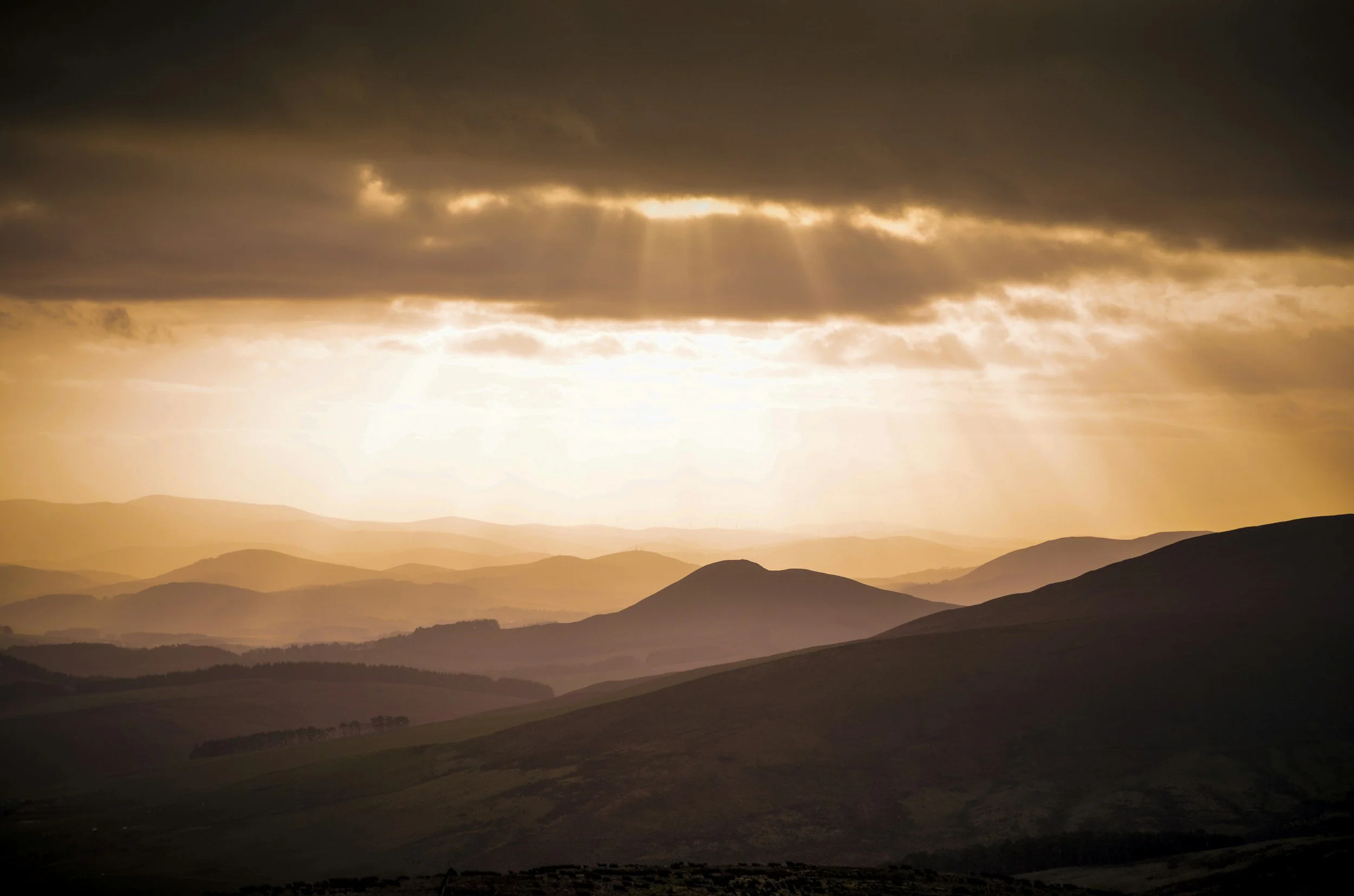 Sunset over rolling hills with dark clouds and rays of sunlight breaking through.