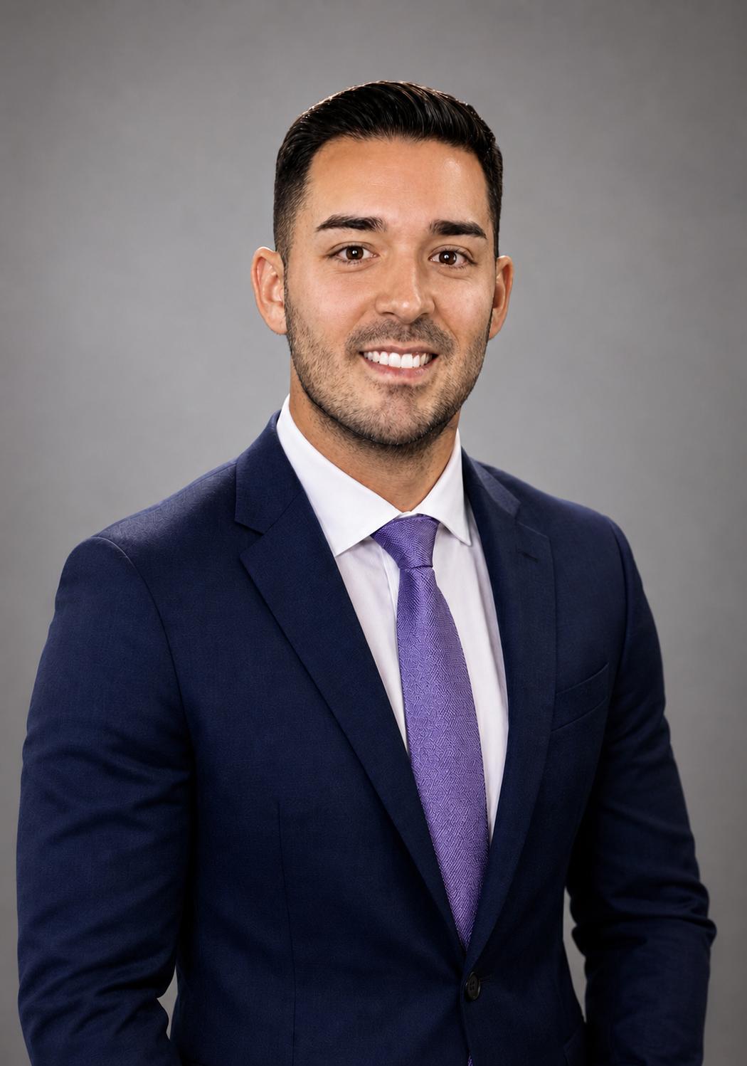 A man in a dark blue suit with a white shirt and purple tie, smiling against a plain gray background.