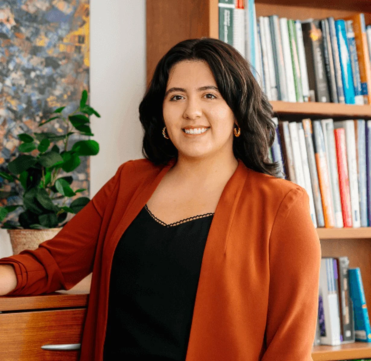 A woman with shoulder-length dark hair smiling in an office or library, wearing gold hoop earrings and a rust-colored blazer over a black top, with a bookshelf filled with books and a green plant in the background.