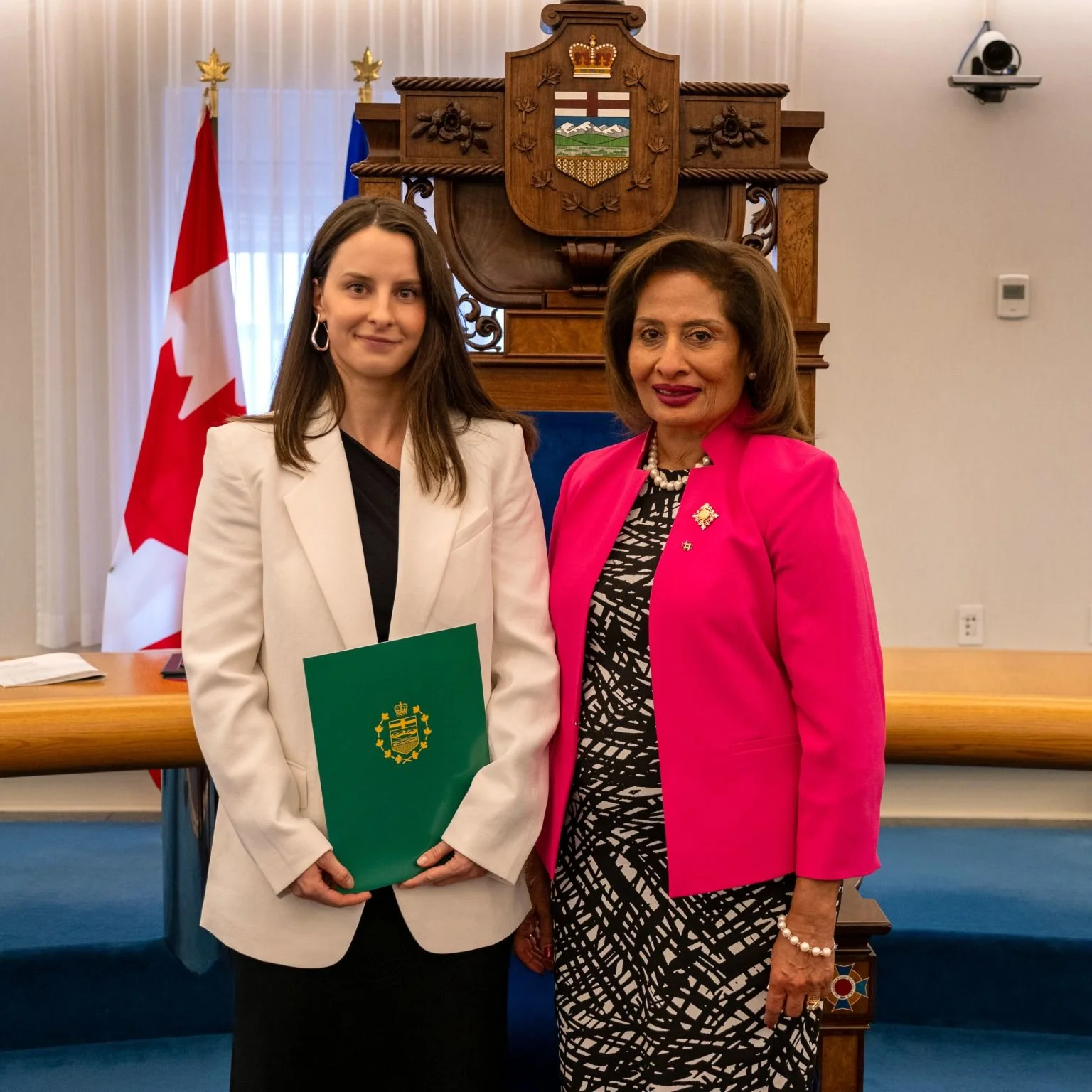 Two women standing in an official setting, with flags and a wooden emblem behind them, one holding a green folder with a crest, both smiling.