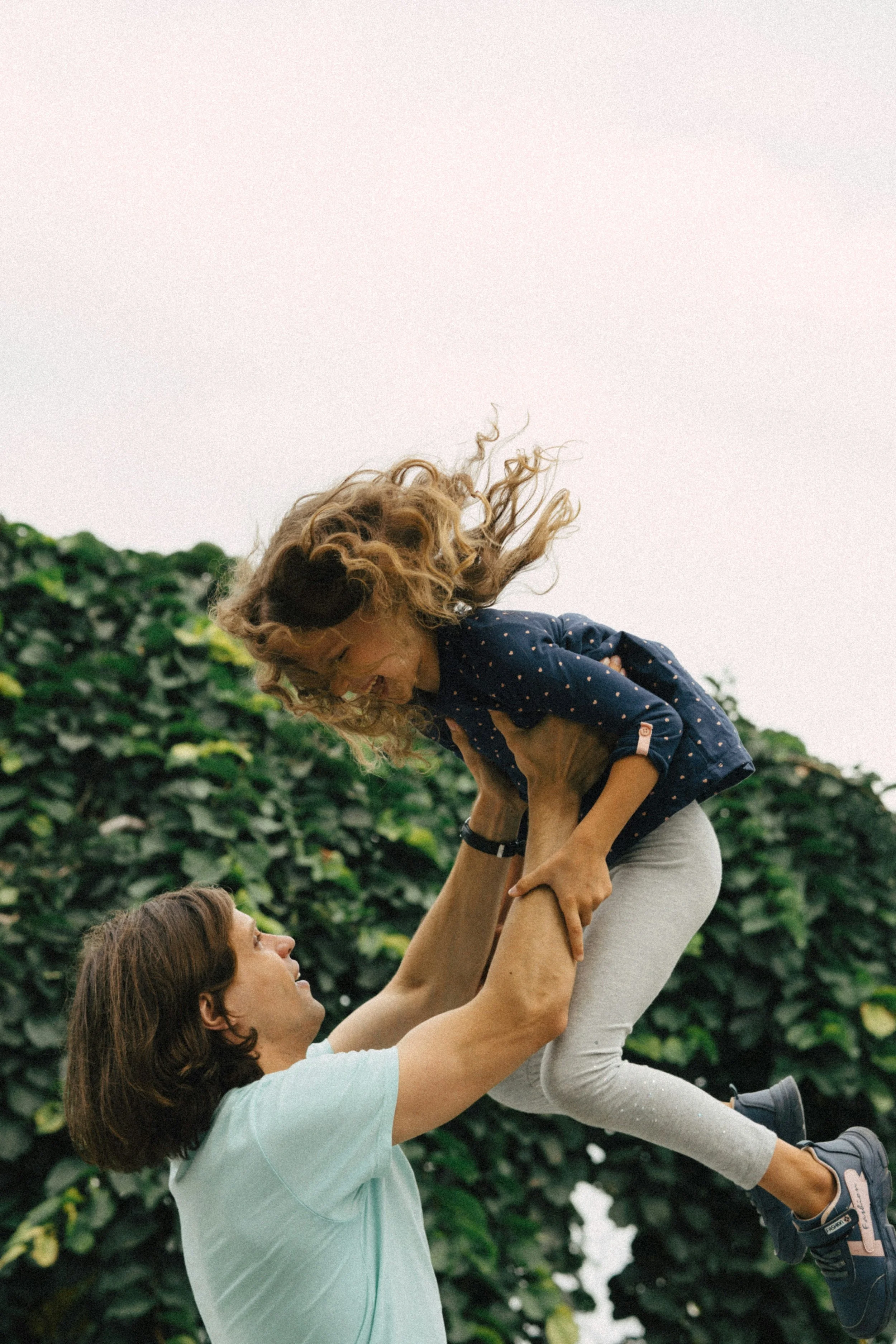 Two women playing with a young girl outdoors, one woman holding the girl in the air, all smiling and having fun. Green bushes in the background.