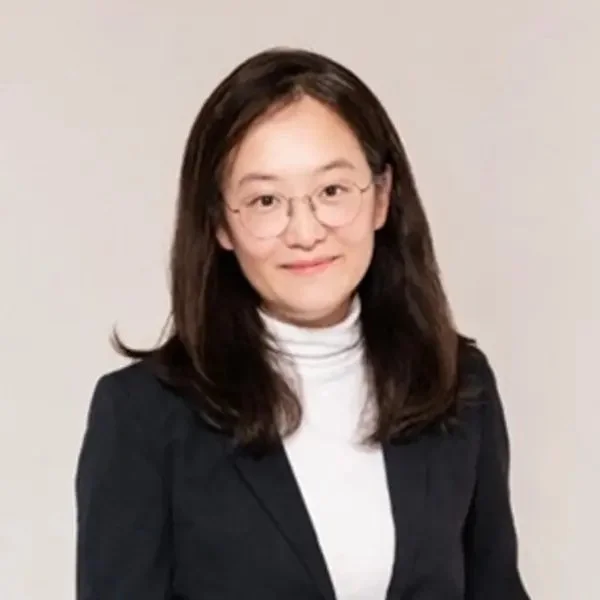 A woman with shoulder-length dark hair and glasses, wearing a white turtleneck and a black blazer, posing against a plain light-colored background.