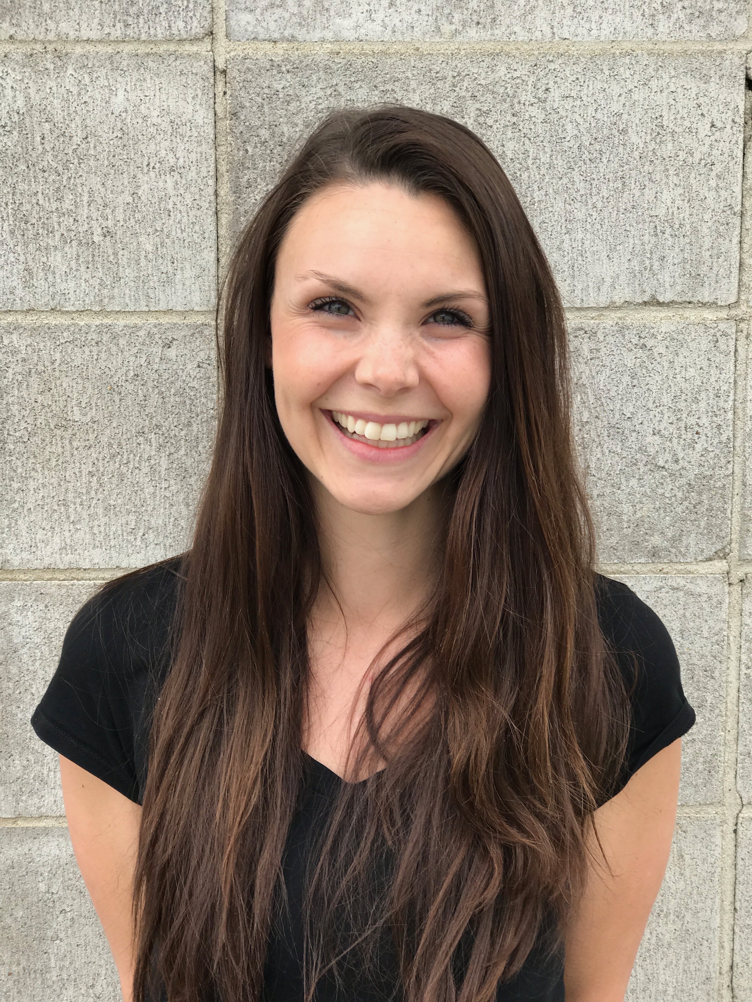 A young woman with long brown hair and blue eyes smiling, standing in front of a light-colored brick wall.