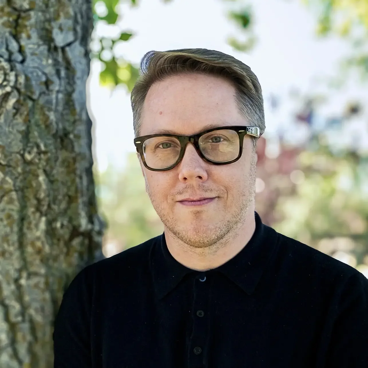 A young man with light skin, short brown hair, and glasses standing outdoors by a tree, smiling at the camera. He is wearing a black shirt and the background is blurred with green foliage.