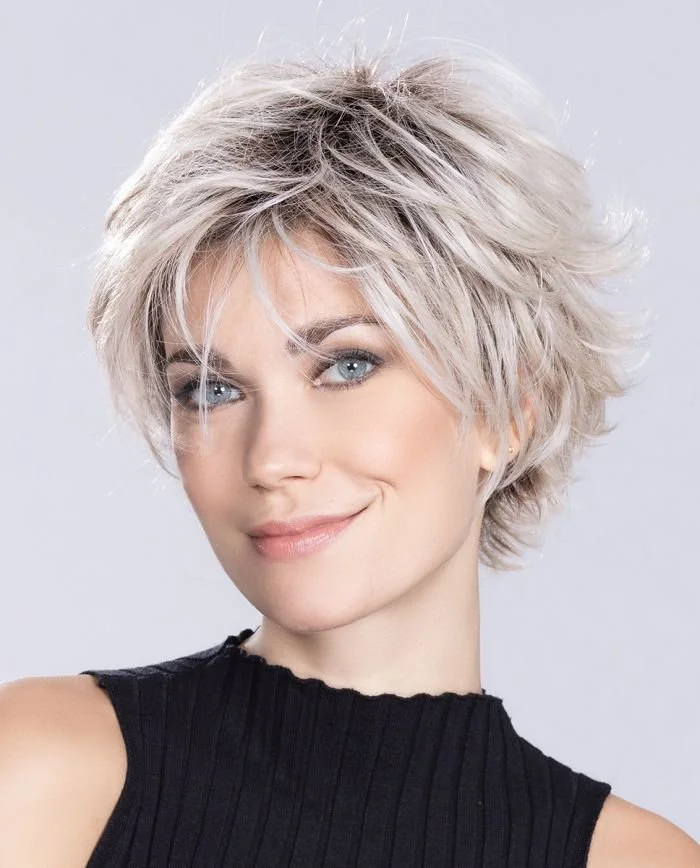 Portrait d'une femme aux cheveux courts, blonds et bouclés, souriant, portant un haut noir sans manches sur un fond gris clair.