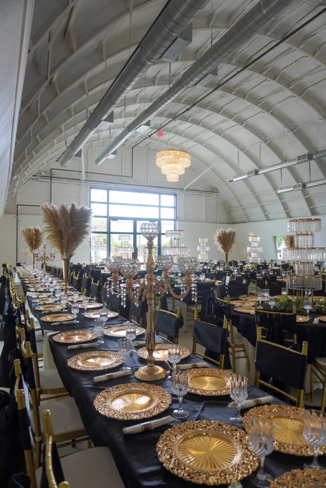 Elegant banquet hall with black tablecloths, gold chargers, crystal glassware, tall gold candle holders with pampas grass, and black chairs with gold accents, decorated for a formal event.
