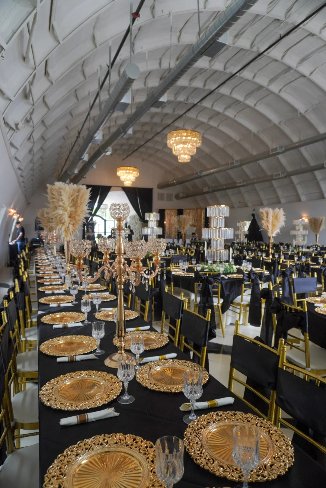 Elegant banquet hall decorated with black tablecloths, gold chargers, crystal glassware, tall gold candelabras with crystal accents, and tall arrangements of pampas grass. The ceiling features large chandeliers and exposed ductwork. Black and gold chairs surround the tables, and there is a stage at the back with black curtains and gold decorations.
