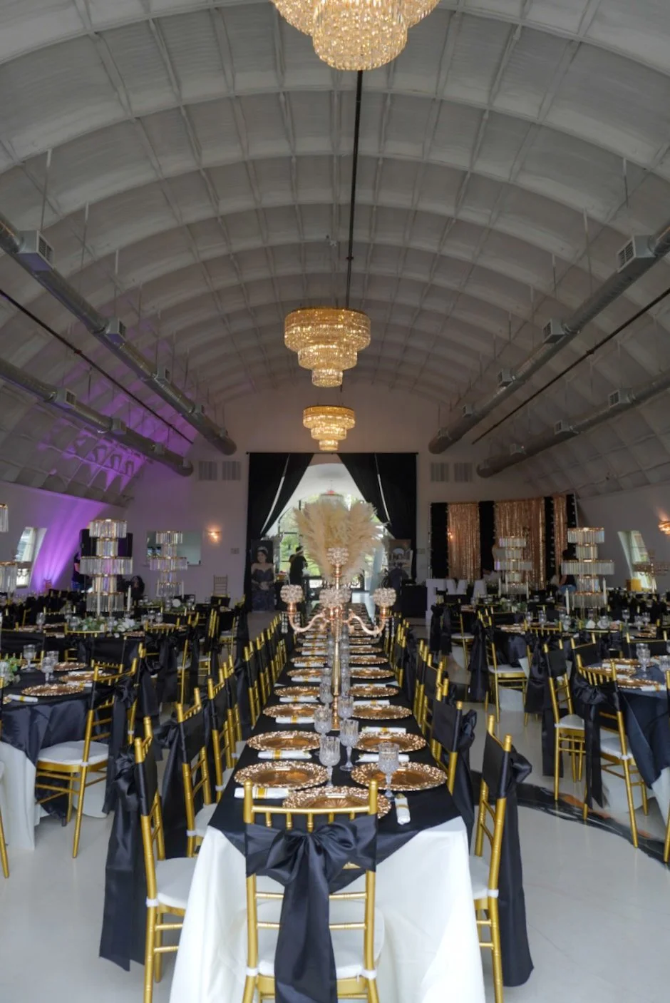 Elegant banquet hall decorated with black, gold, and white theme including gold chairs with black sashes, black tablecloths, and gold accents. The ceiling features large, gold chandeliers. The tables are set with gold chargers, glassware, and centerp