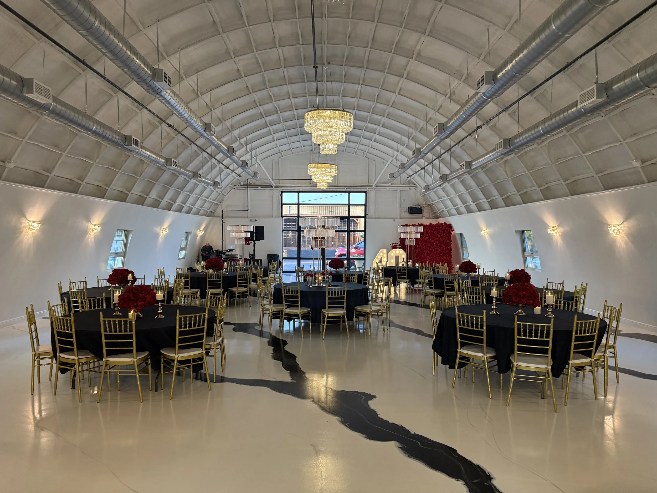 Ballroom decorated for an event with round tables covered with black tablecloths, gold chairs, and red floral centerpieces. Large chandeliers hang from a high arched ceiling with exposed ductwork. There is a flower wall and large windows at the front
