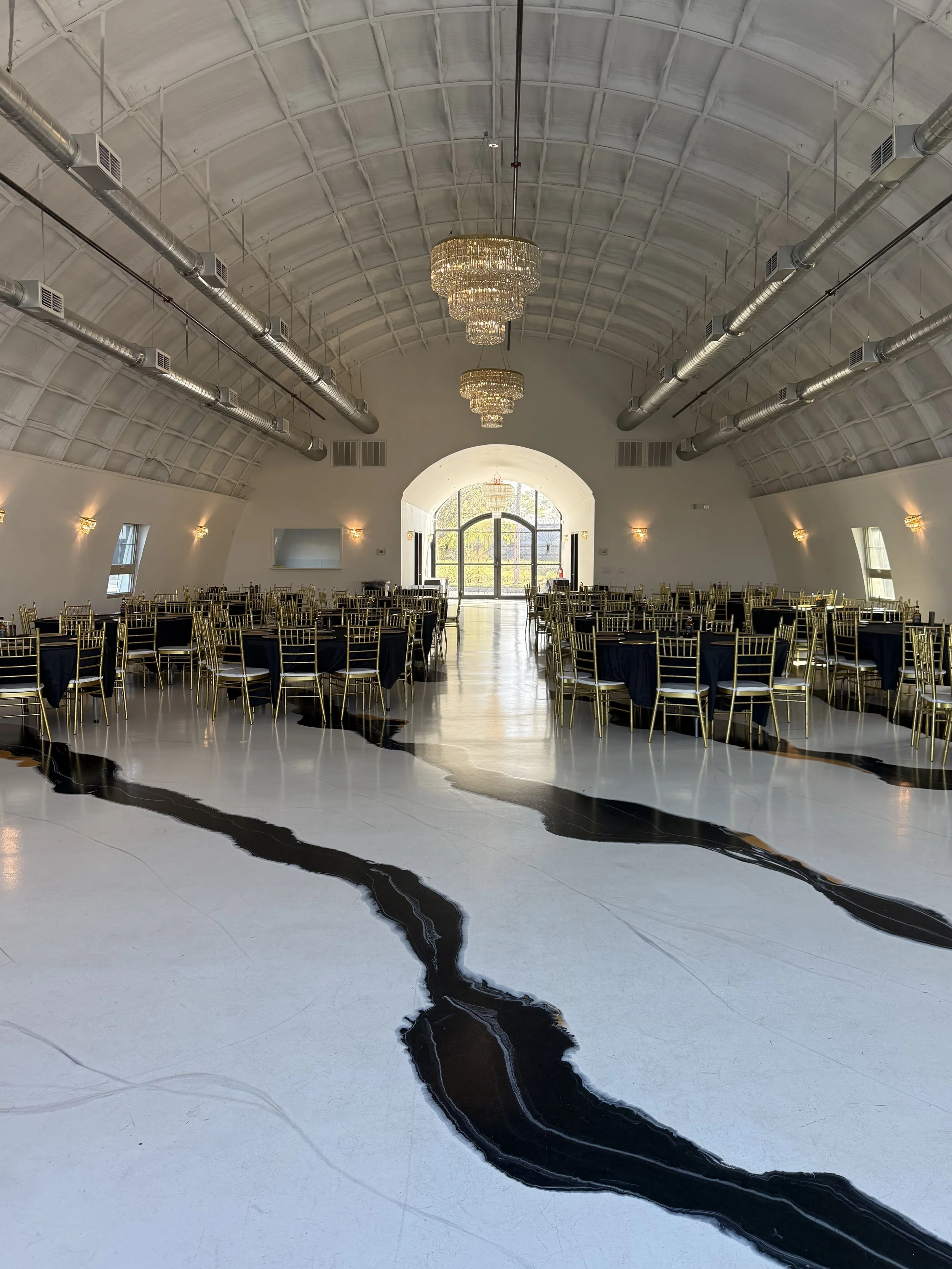 A large banquet hall with high arched ceiling, chandeliers, and gold chairs around decorated tables, with a black and white marble floor and natural light coming through windows and the main door.