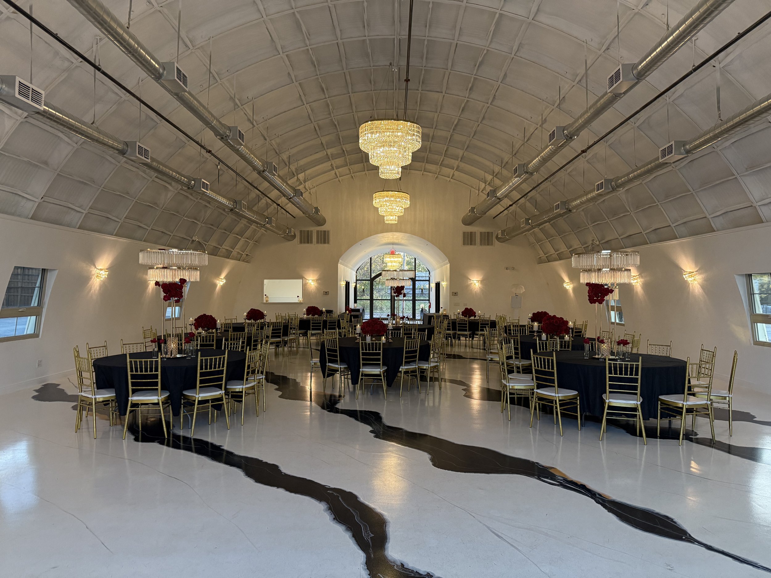 Elegant banquet hall with high vaulted ceiling, multiple chandeliers, round tables with black tablecloths, gold chairs, and red floral centerpieces. Large arched window at the back allows natural light.