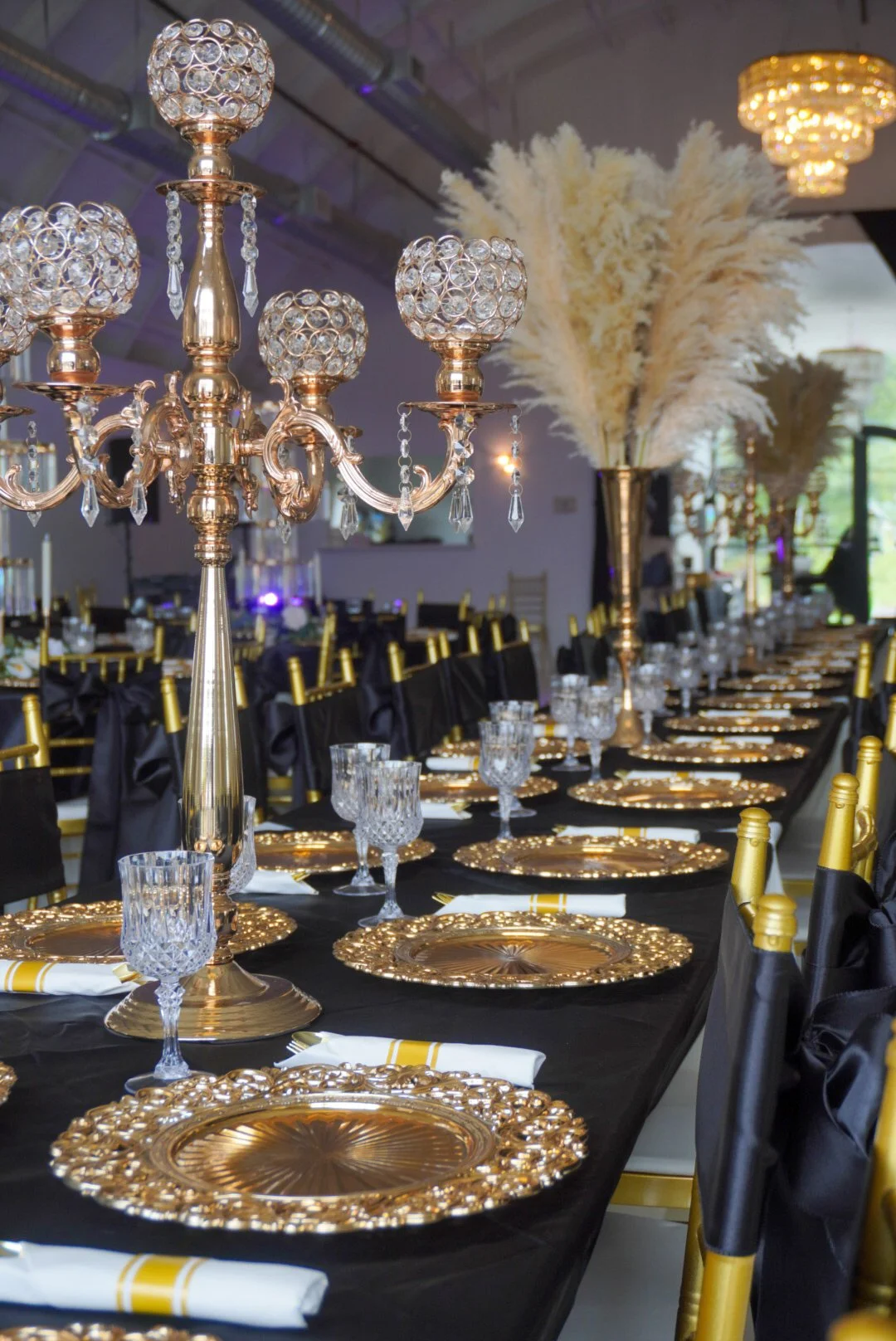 Elegant banquet table setting with gold and black decor, featuring ornate gold chargers, crystal glasses, tall gold candelabras with crystal accents, black napkins with gold rings, and large white floral arrangements, in a decorated event hall with d