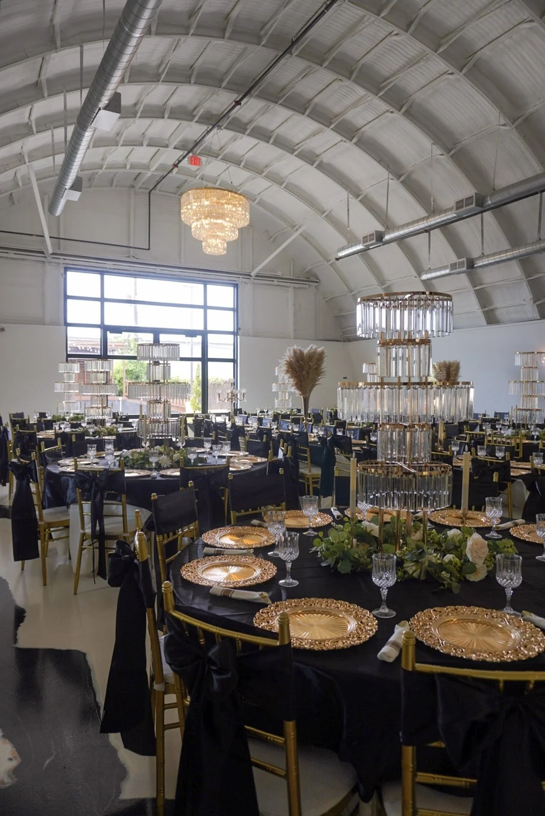 Elegant event space with black tables, gold chairs, black tablecloths, and gold chargers. Centerpieces feature tall glass candle holders with crystal accents and pampas grass. Crystal chandelier and modern lighting fixtures hang above, large front wi