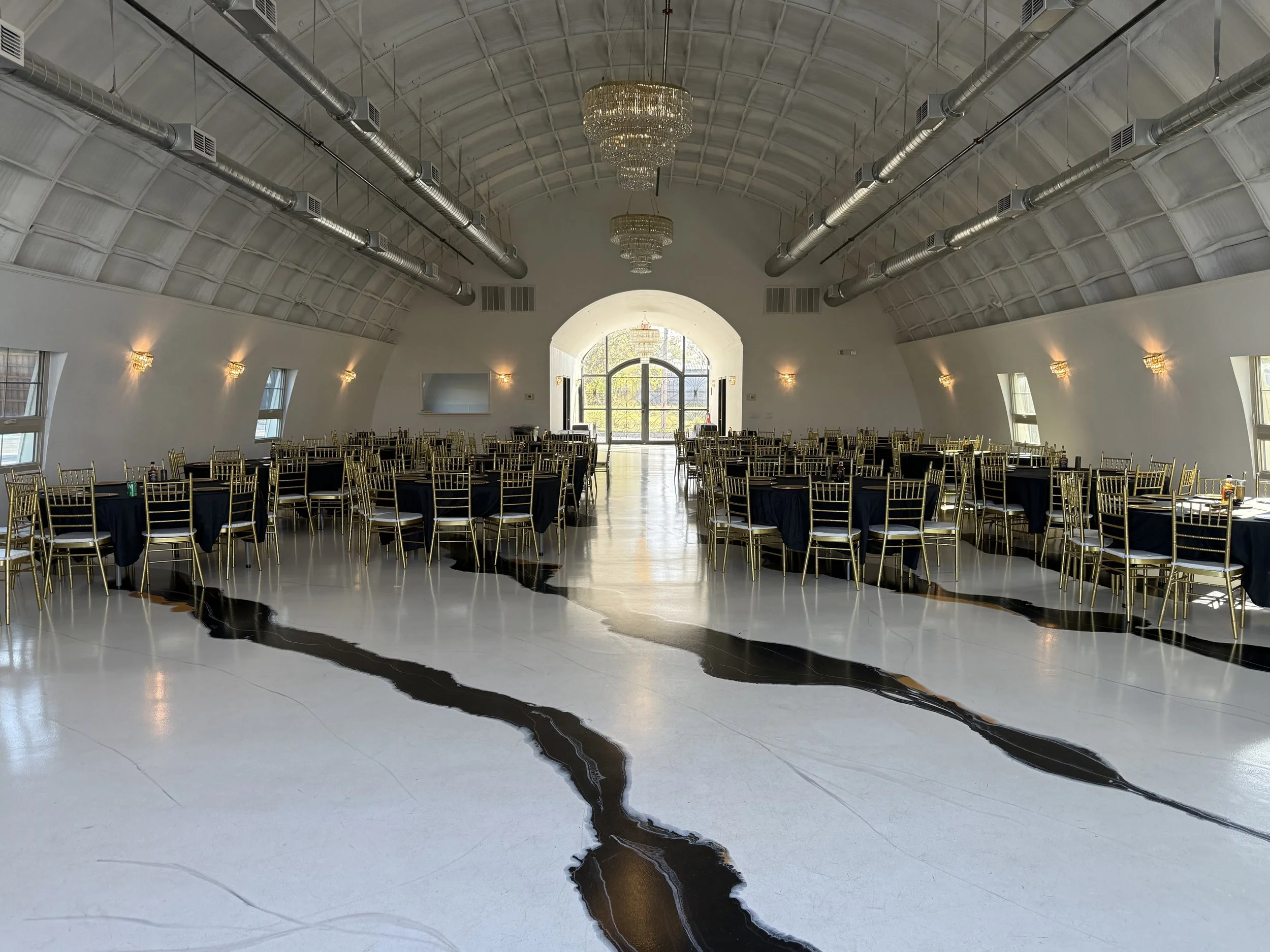A large banquet hall with a high, arched ceiling featuring chandeliers and ductwork. Elegantly set tables with black tablecloths and gold chairs are arranged along the sides, leaving a clear walkway in the center. Natural light streams in through win