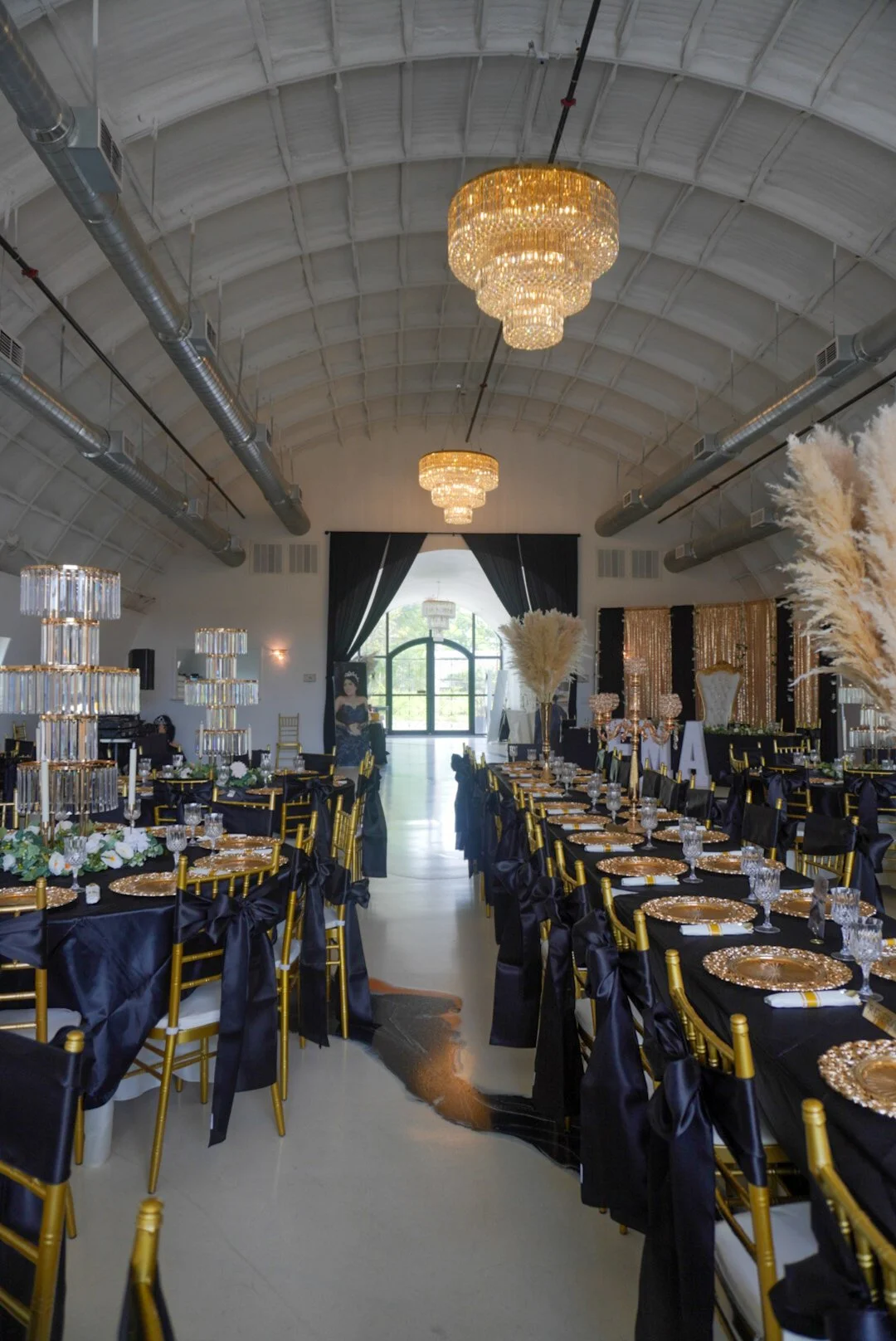 Elegant banquet hall with gold Chiavari chairs, black tablecloths, and gold chargers, decorated with tall floral centerpieces and crystal chandeliers hanging from a vaulted ceiling.