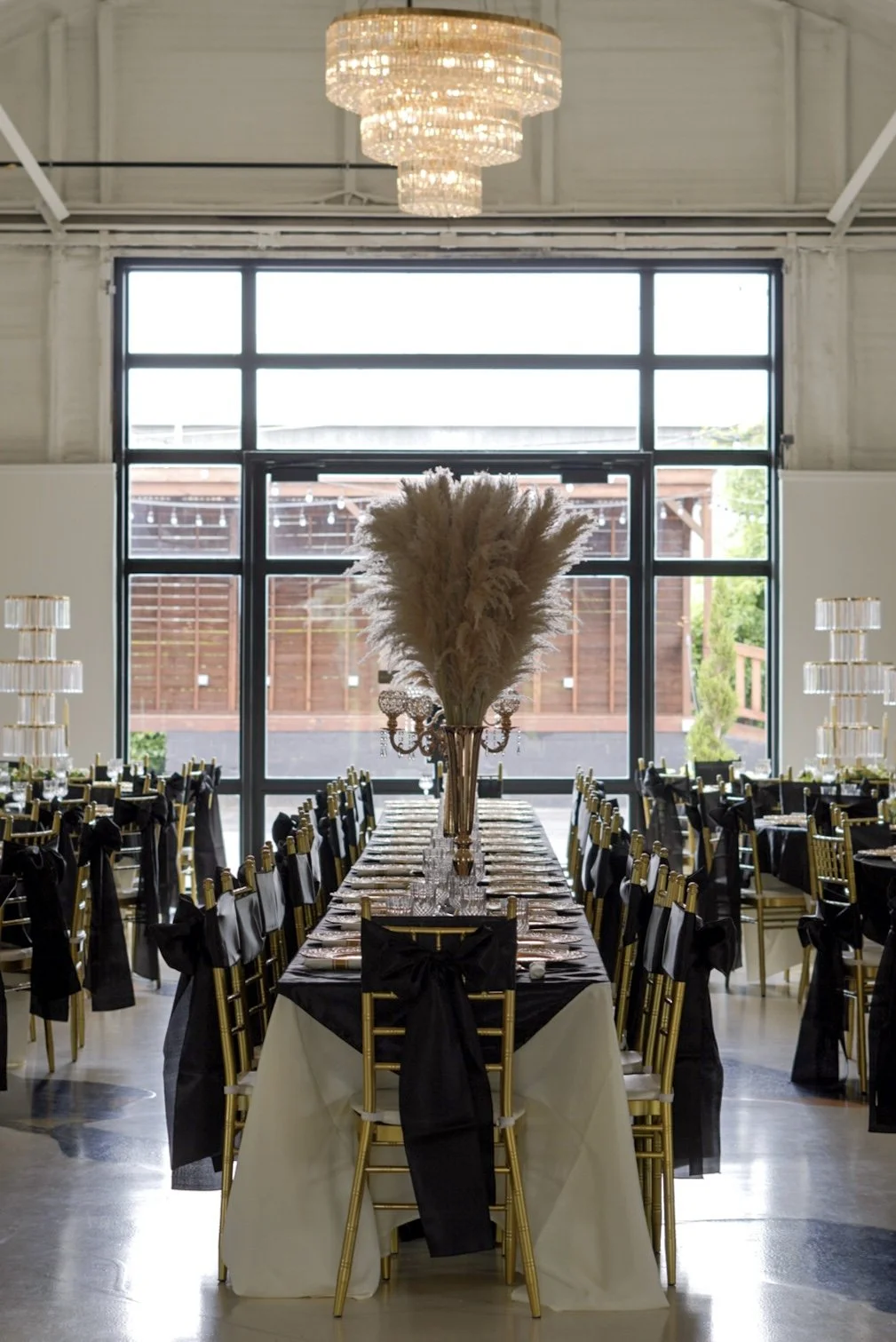 Elegant banquet hall decorated for a formal event, featuring a large central table with gold chairs, black and white tablecloths, and tall vases with pampas grass centerpiece. Crystal chandeliers hanging from the ceiling and a large window with a vie