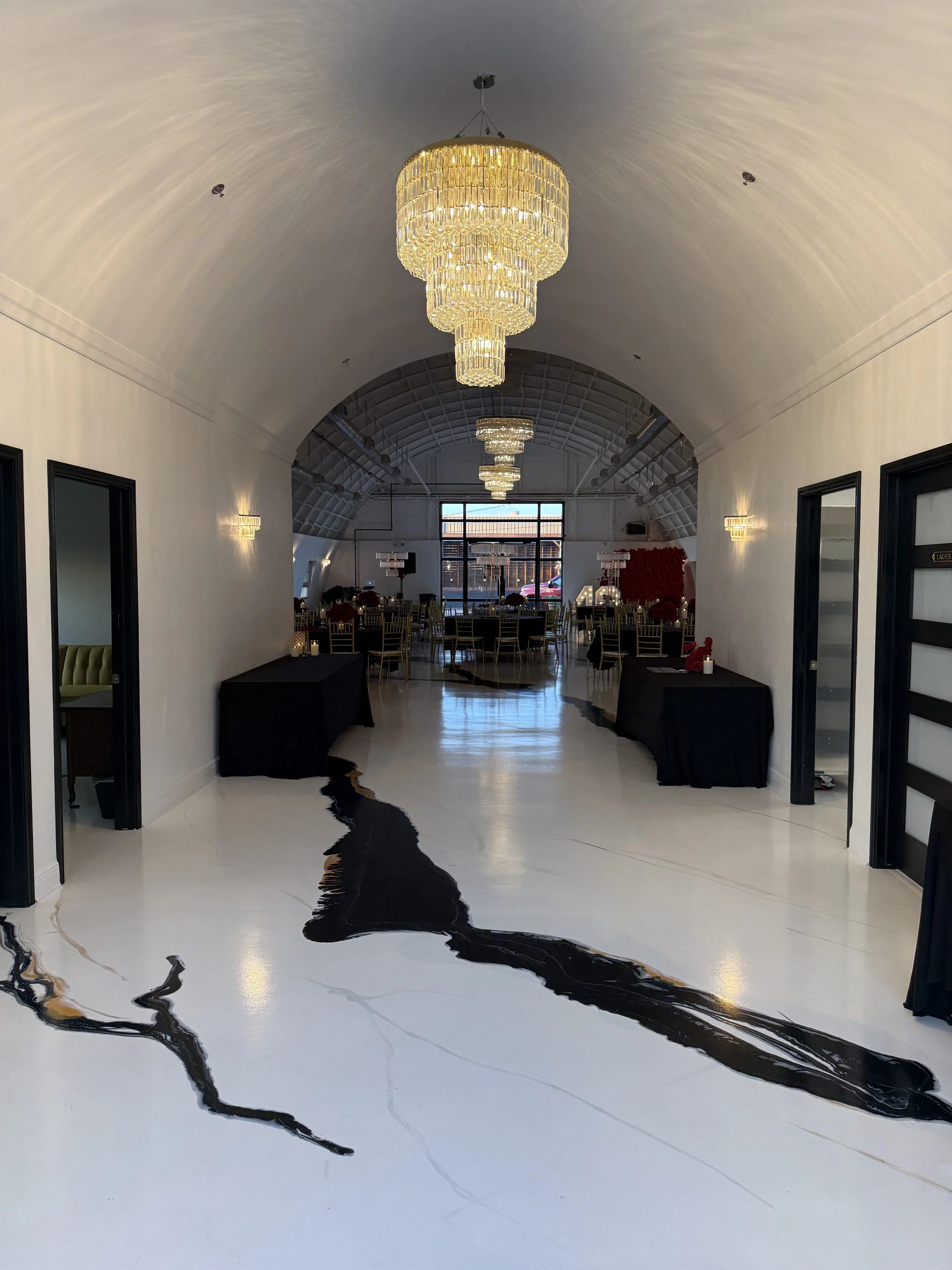 Elegant banquet hall with a large, elaborate chandeliers hanging from a vaulted ceiling. The hall is decorated with round tables, each with chairs and centerpieces. The floor has a black and white swirling pattern that resembles a marble or ink desig