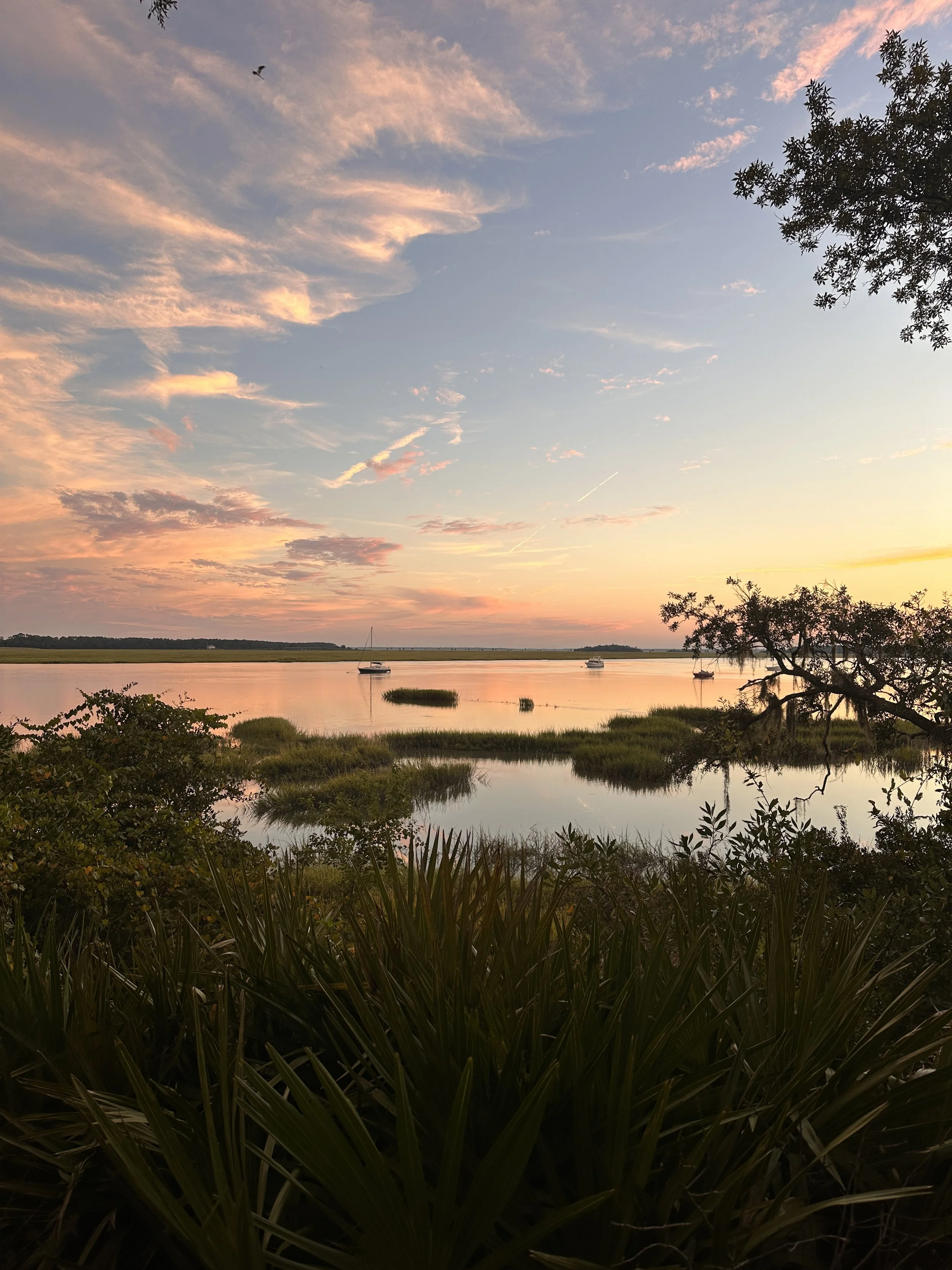 A sunset over Battery Creek with boats anchored near the shore, surrounded by trees and lush vegetation, with colorful clouds in the sky.