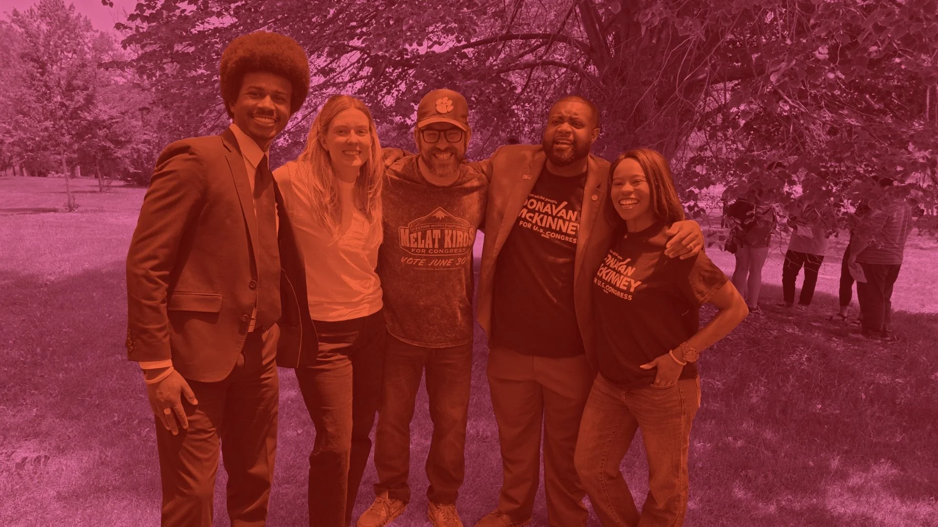 Group of six diverse people standing outdoors on grass, smiling, with trees in the background, some wearing campaign T-shirts, some with their arms around each other.