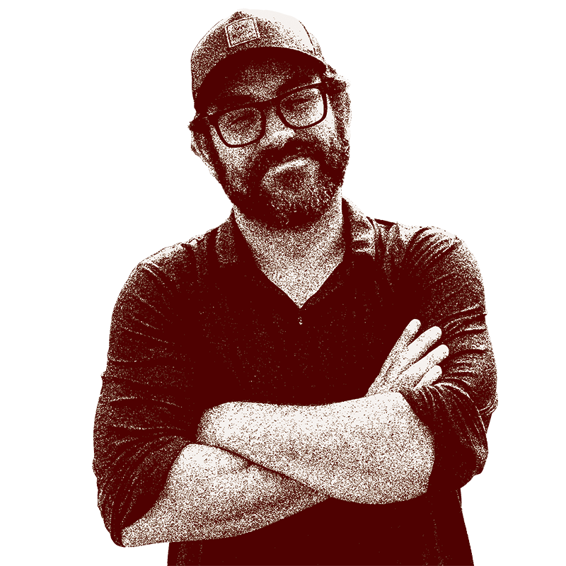 A man with glasses, a beard, and a cap, wearing a dark shirt with rolled-up sleeves, crossing his arms against a black background.