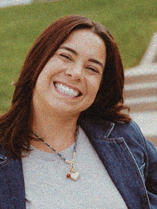 Smiling woman with long auburn hair, wearing a denim jacket and a heart-shaped necklace, outdoors on a grassy area.