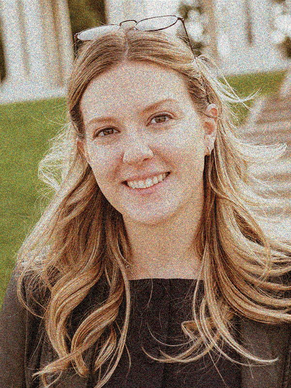 A woman with long, wavy blond hair smiling outdoors, wearing glasses on her head and a black top.