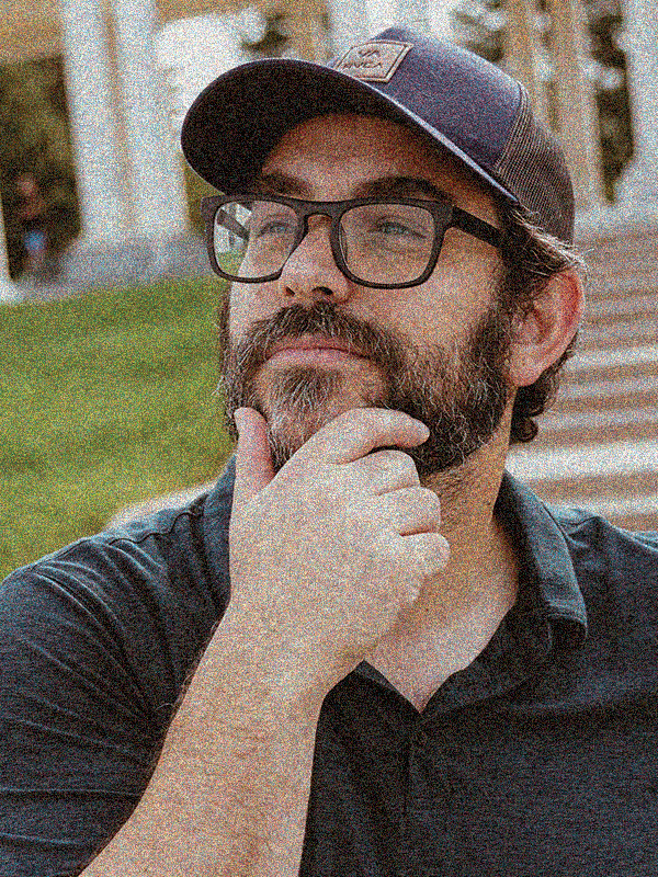 A man with glasses and a beard, wearing a baseball cap, standing outdoors near some steps, thoughtfully resting his chin on his hand.