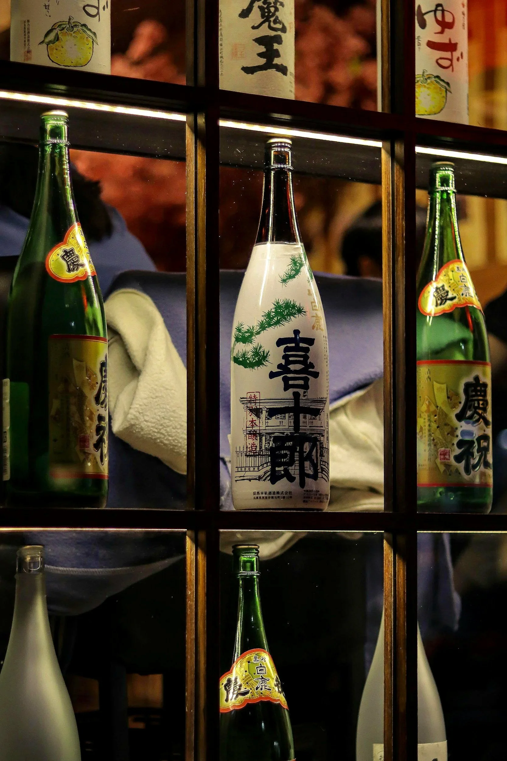 A shelf displaying several bottles of Japanese sake in a restaurant setting. The bottles have traditional labels with Japanese characters and images, and are arranged behind each other, with a person with dark hair and a blue shirt visible in the background.