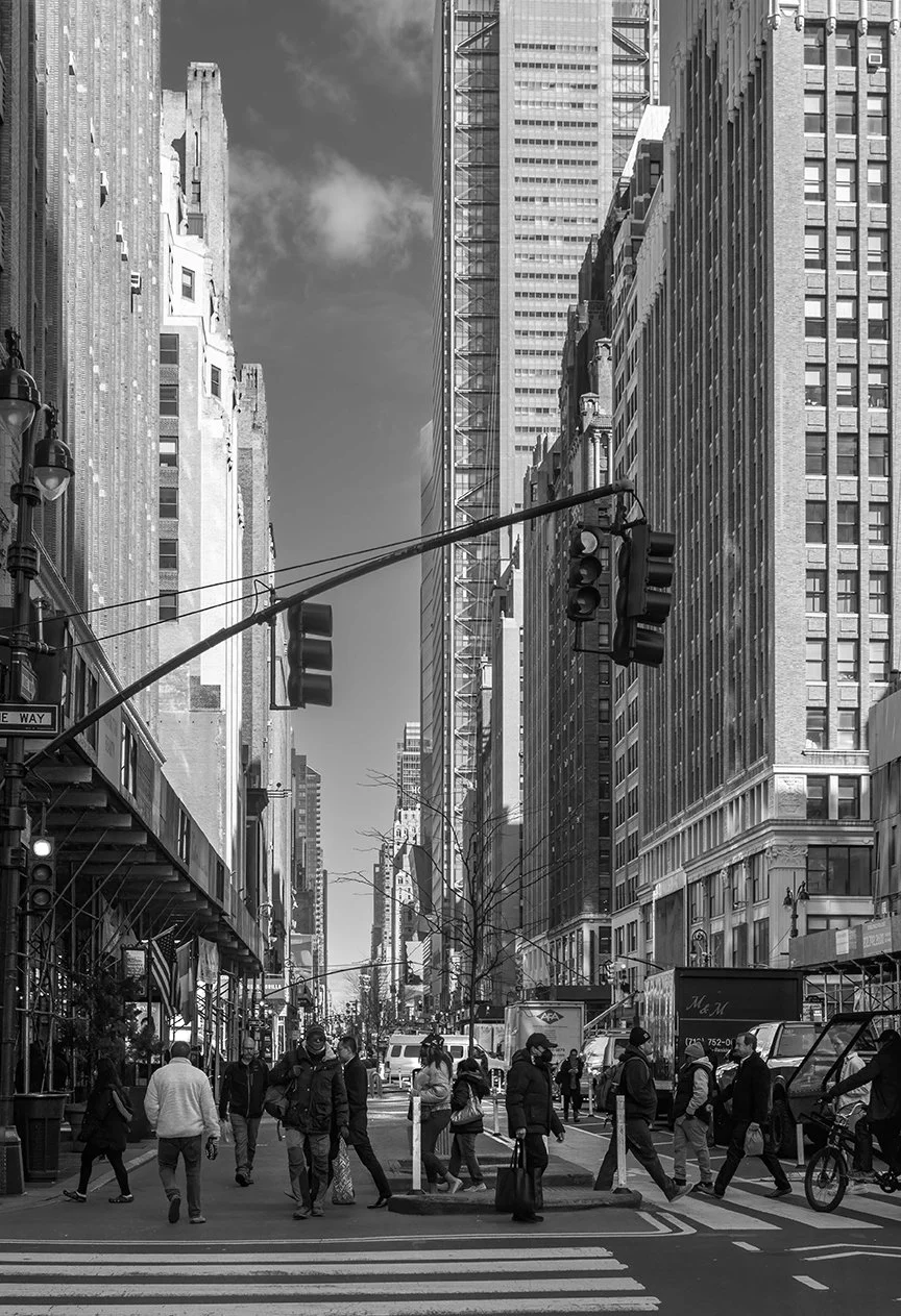 Black and white photo of a busy city street with people crossing the crosswalk, tall skyscrapers on both sides, and traffic lights overhead.
