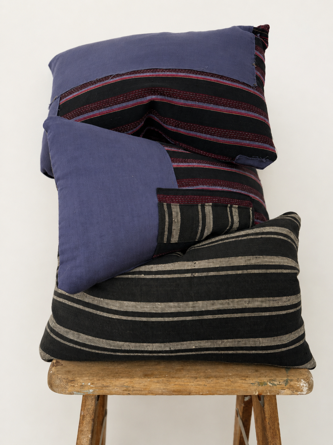 Three stacked decorative pillows with striped patterns, placed on a wooden stool against a plain background.