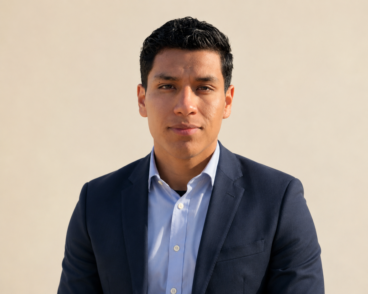A young man in a navy blazer and white shirt, standing against a plain light-colored background, looking directly at the camera.