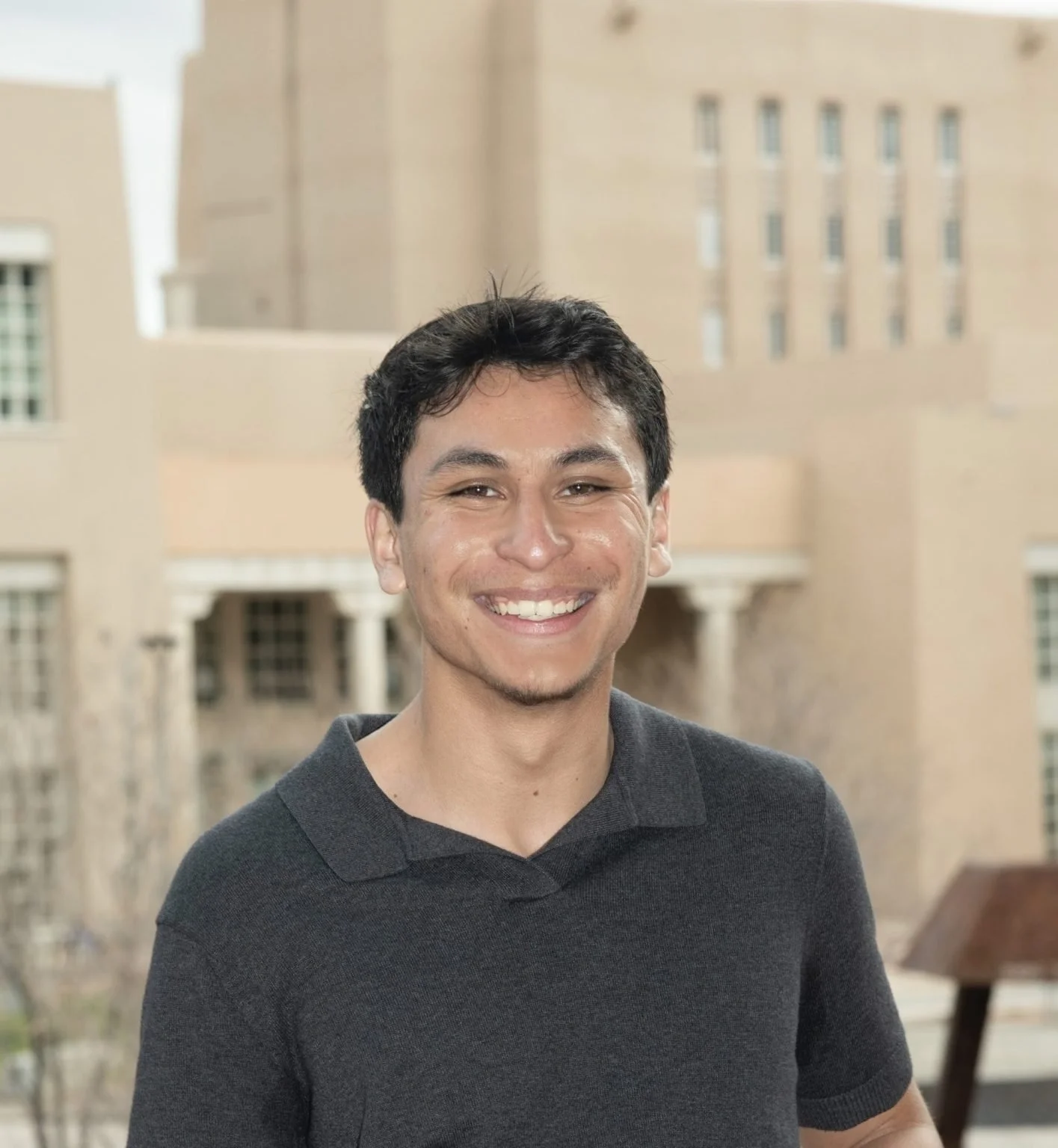 Young man with short black hair and a mustache smiling outdoors in front of a beige building with multiple windows.