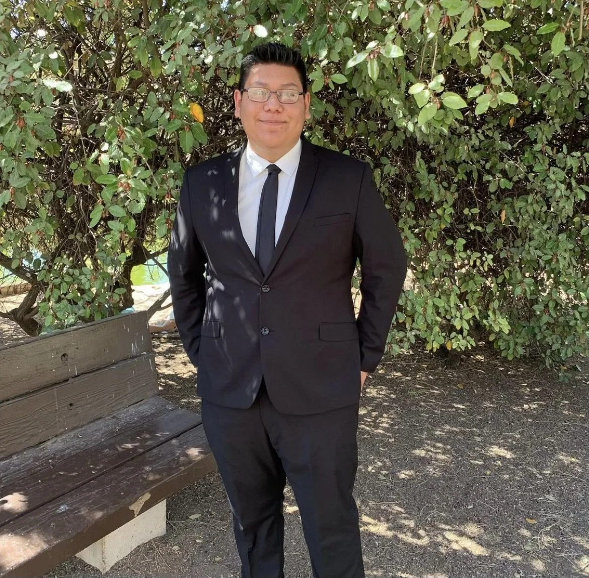A young man in a black suit, white shirt, and black tie standing outdoors in front of green leafy bushes. He is smiling and has glasses. There is a wooden park bench to his left.
