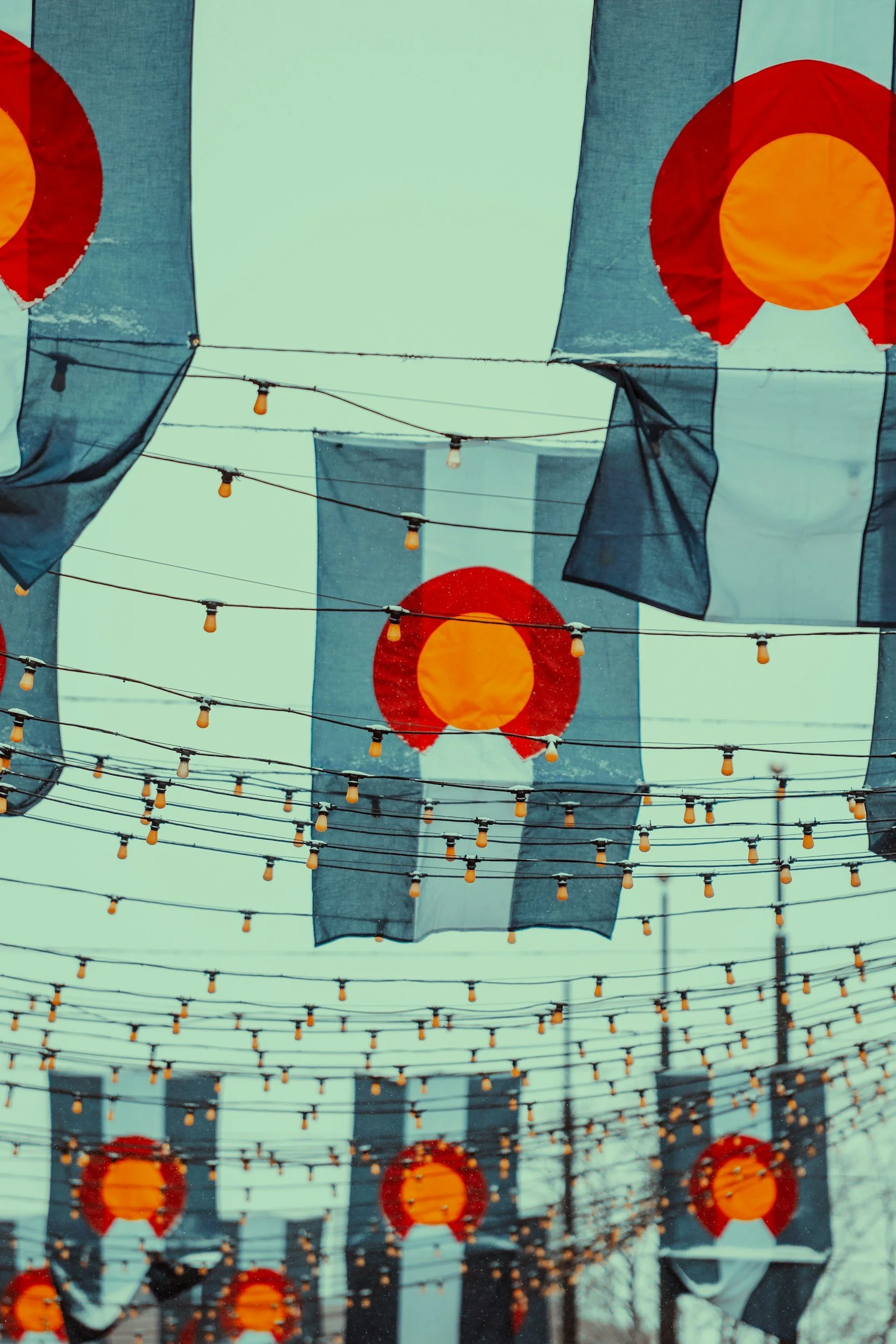 Festive outdoor scene with hanging paper lanterns and string lights in an urban setting, with modern high-rise buildings in the background.
