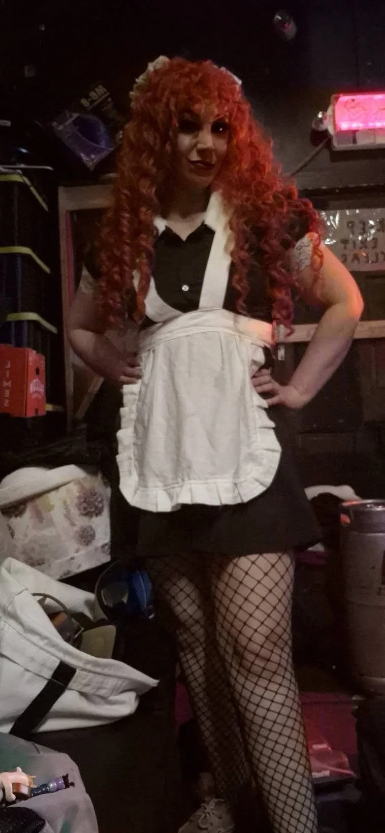Person with curly red hair dressed in a vampiress costume, wearing fishnet stockings and a black dress with a white apron, in a dimly lit room with various objects and bottles.