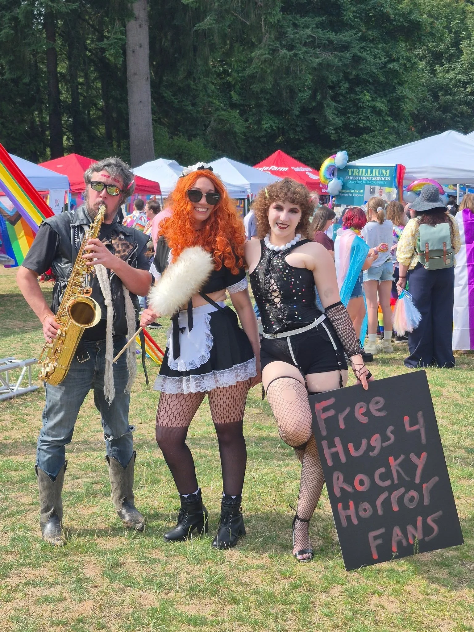 Three women in gothic and punk style clothing posing at an outdoor event, with tents and a crowd in the background. One woman has curly hair and is holding a sign, another has vibrant red hair, and the third is playing a saxophone.