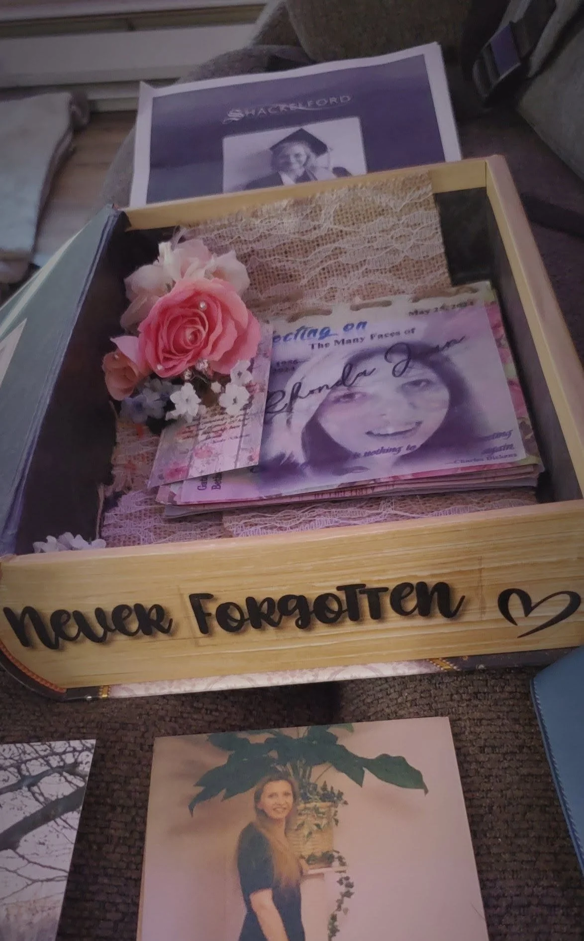 A wooden box with the words "never forgotten" written on it, filled with pink flowers, a photo of a young girl, and a newspaper clipping. There is a school photo of a girl in graduation cap and gown in the background.